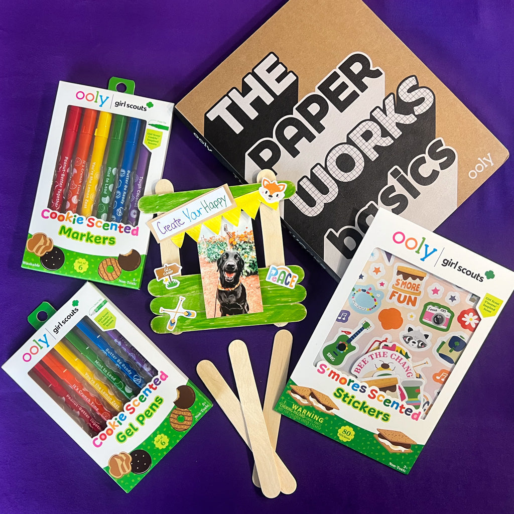 Completed popsicle stick picture frame decorated with OOLY x Girl Scouts cookie-scented markers, gel pens, and s’mores-scented stickers, surrounded by the craft supplies used.