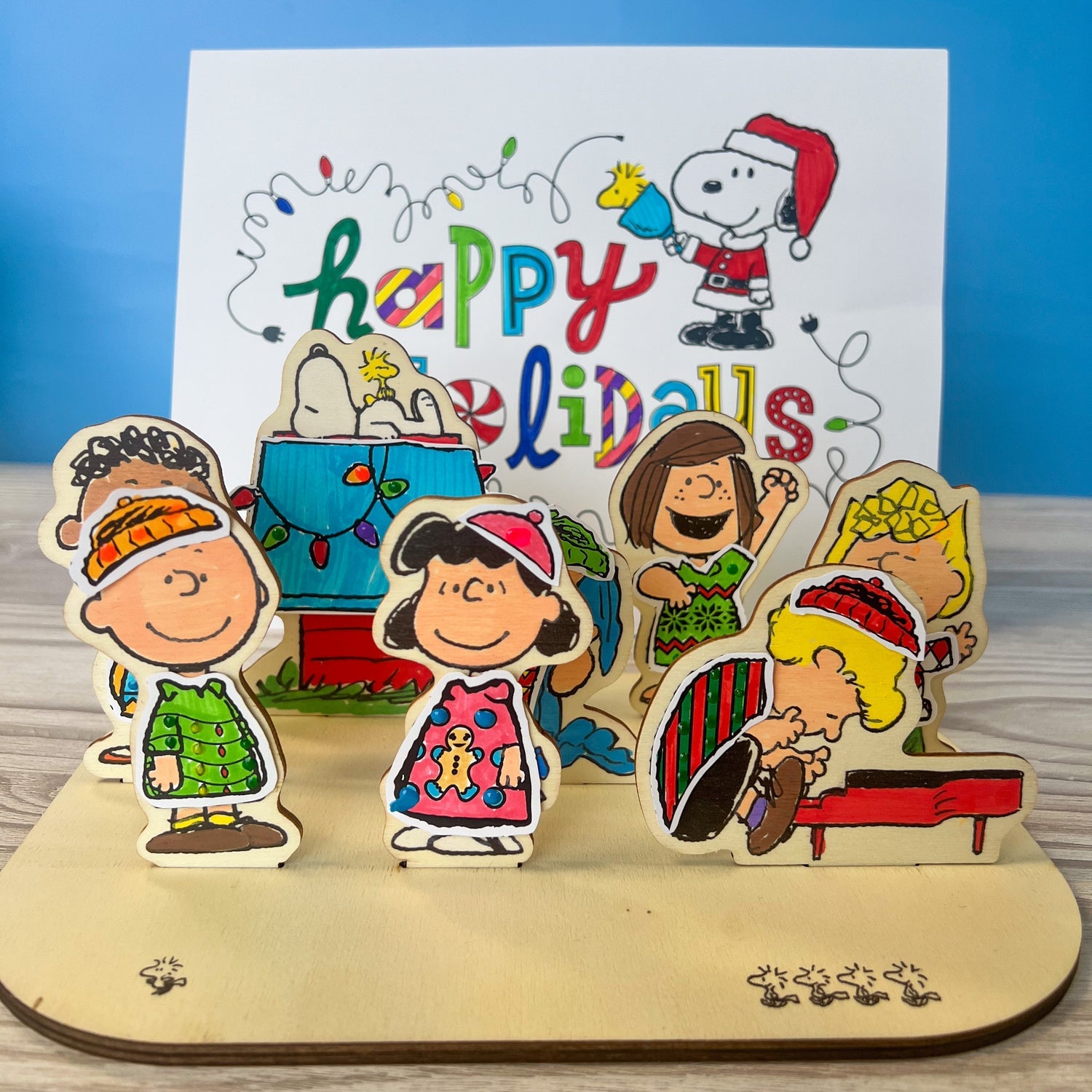 Completed OOLY x Peanuts wooden holiday scene craft featuring Snoopy and friends in colorful sweaters colored with OOLY markers.