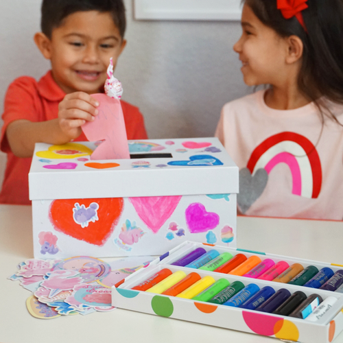 Two kids smiling while dropping Valentine’s cards into their decorated Keepsake Valentine Box, featuring hearts and stickers made with OOLY supplies.