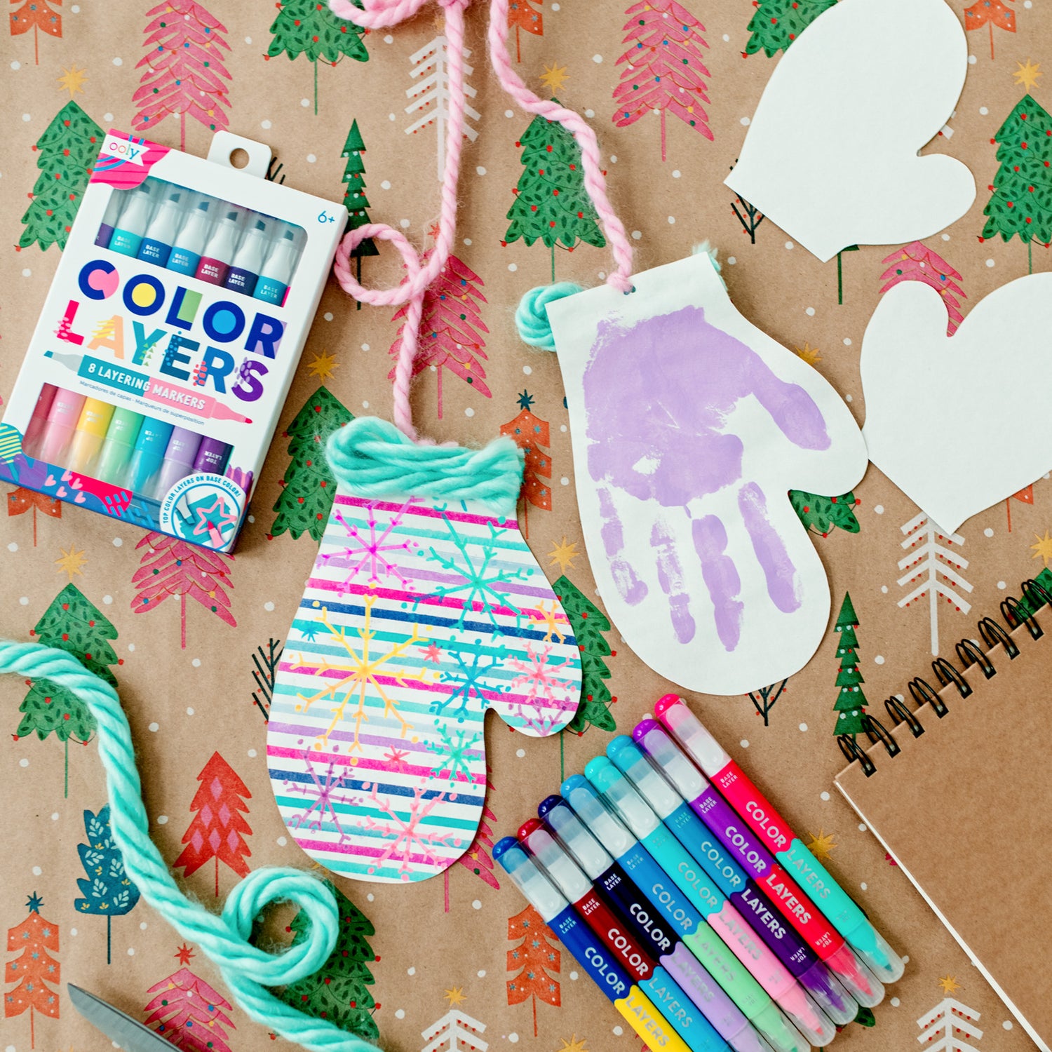 Two paper mittens connected by pink yarn, with an OOLY White DIY Cover Sketchbook and two packages of Ooly Color Layers Double-Ended Layering Markers placed against a backdrop featuring red, pink, and green Christmas trees. Ooly Craft Blog.