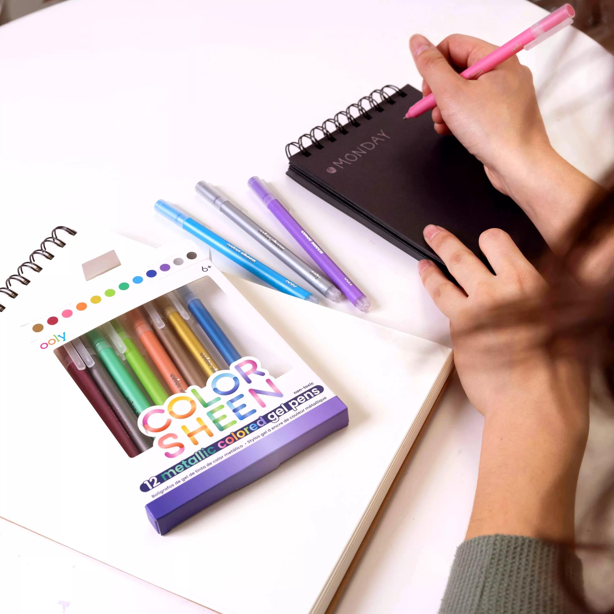 Person writing on a black sketchbook with Color Sheen Metallic Colored Gel Pens