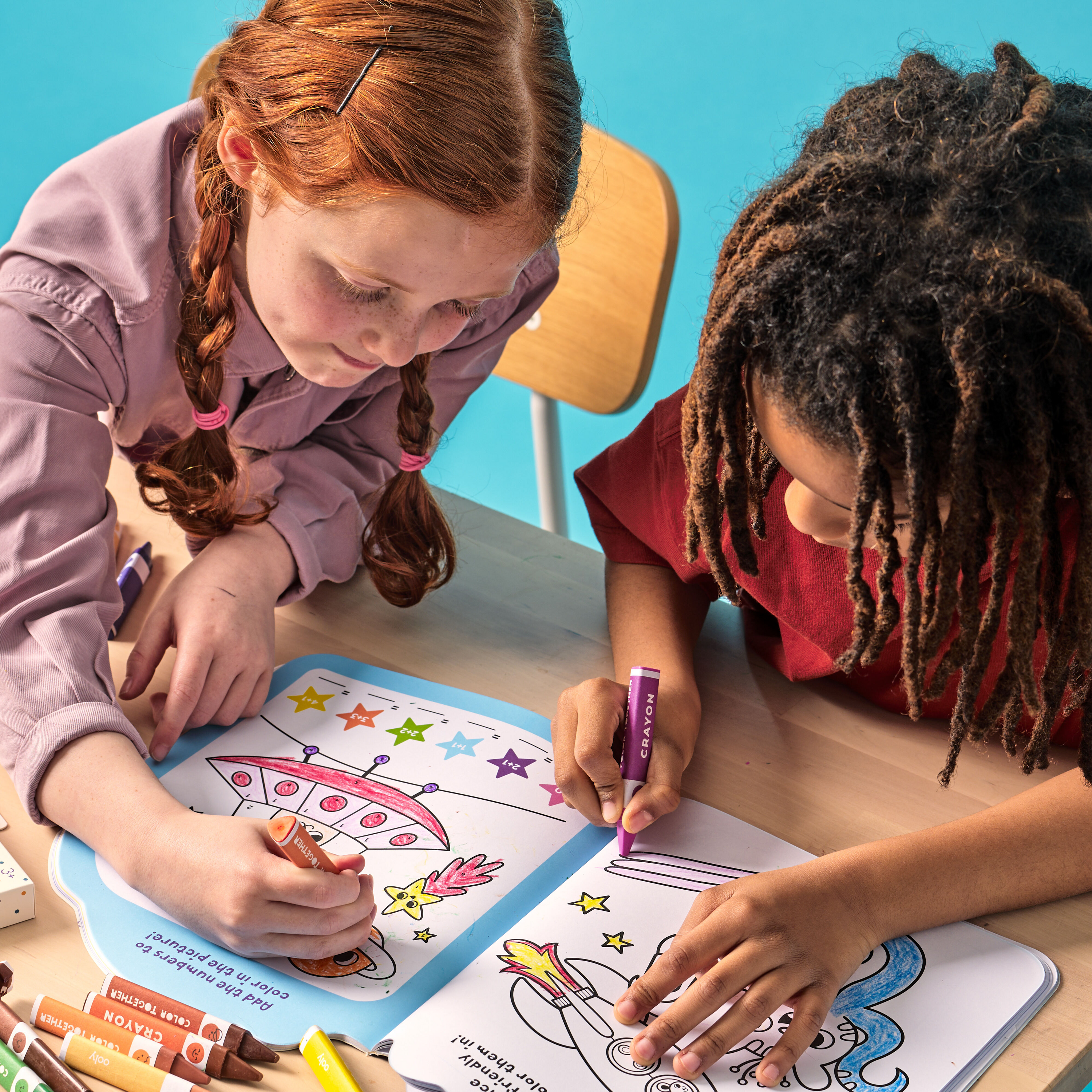 Two kids coloring in activity book with OOLY Color Together Triangular Crayons on a wooden desk