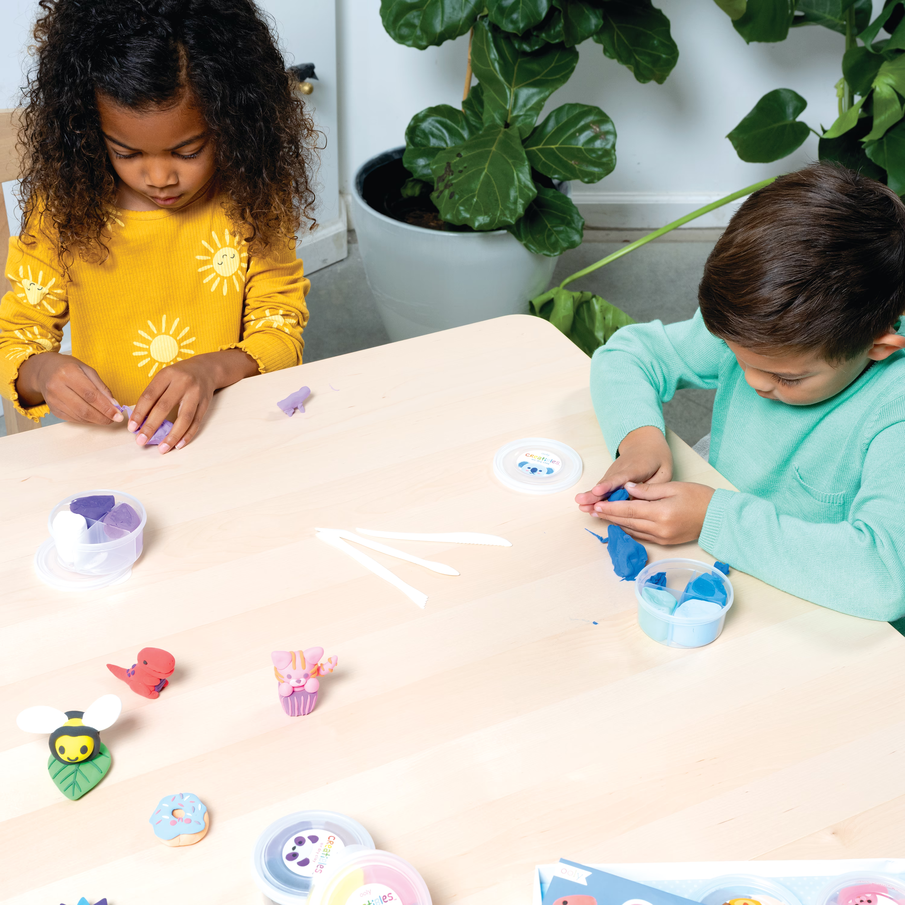 Two kids creating clay sculptures on table top with OOLY Creatibles Deluxe Air Dry Clay Kit