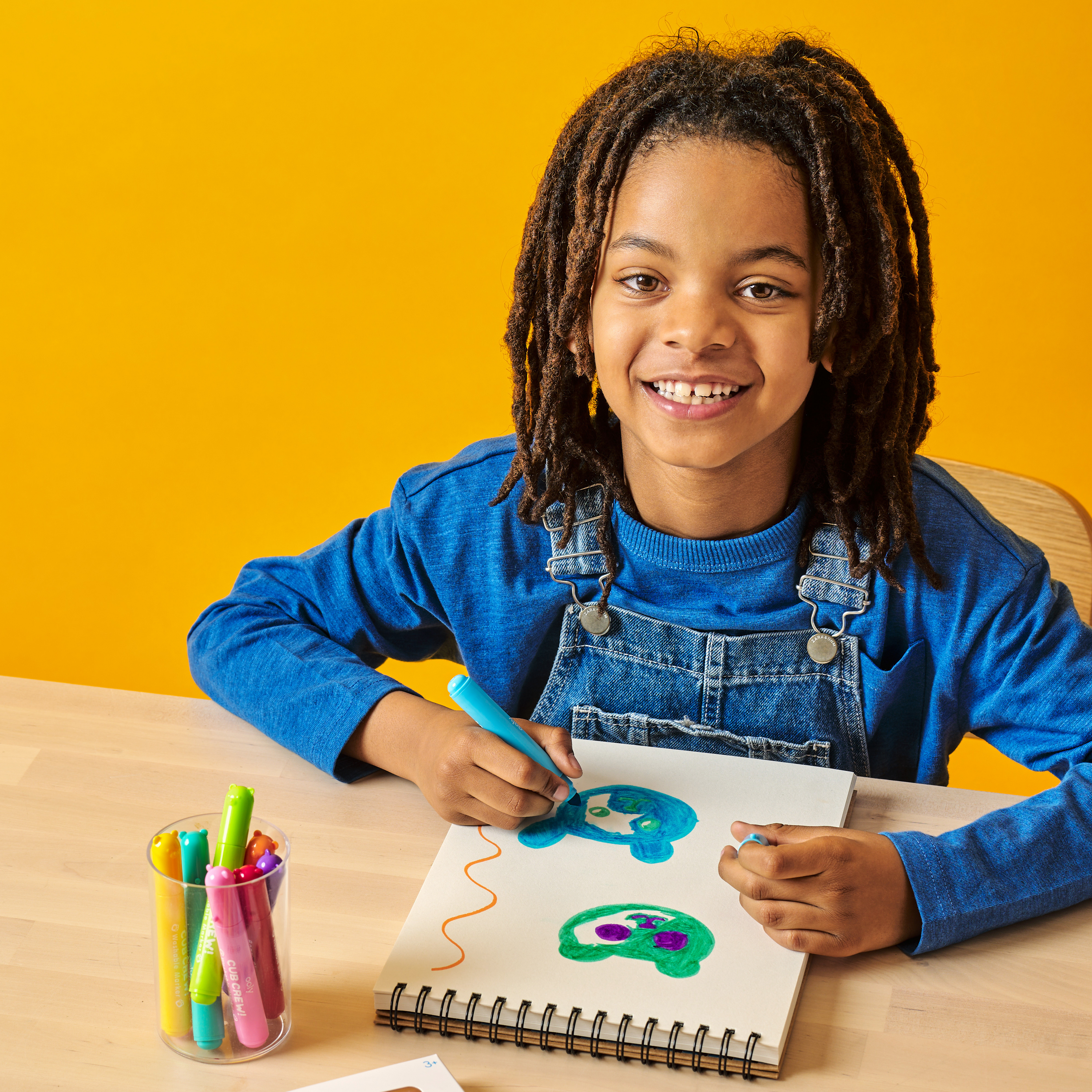Kid smiling while coloring bears in a sketchbook with OOLY Cub Crew Washable Markers on a wooden table