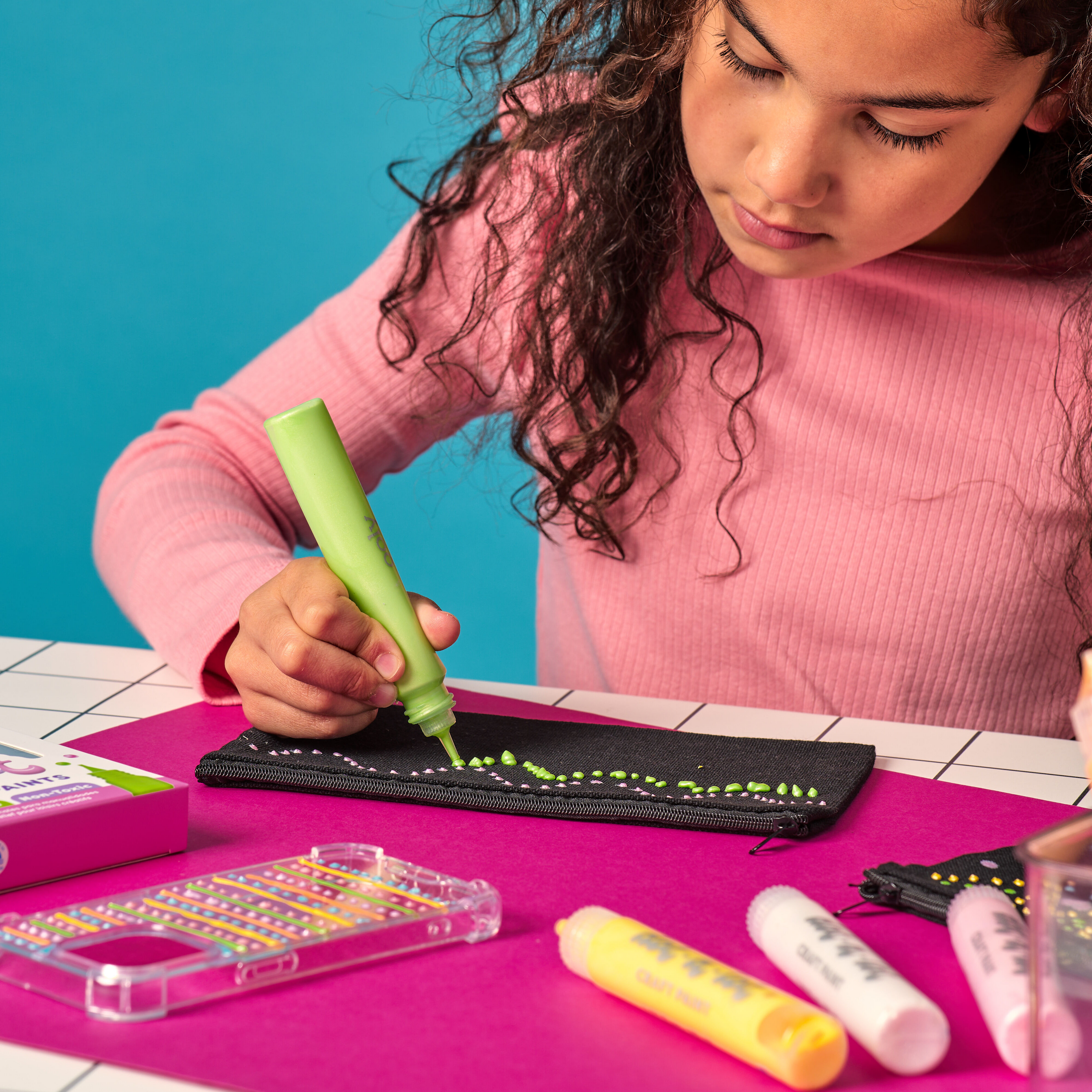 Girl using a green OOLY Dot-a-Lot 3D Dimensional Craft Paint stick on a black pouch with colorful paint on a pink surface.
