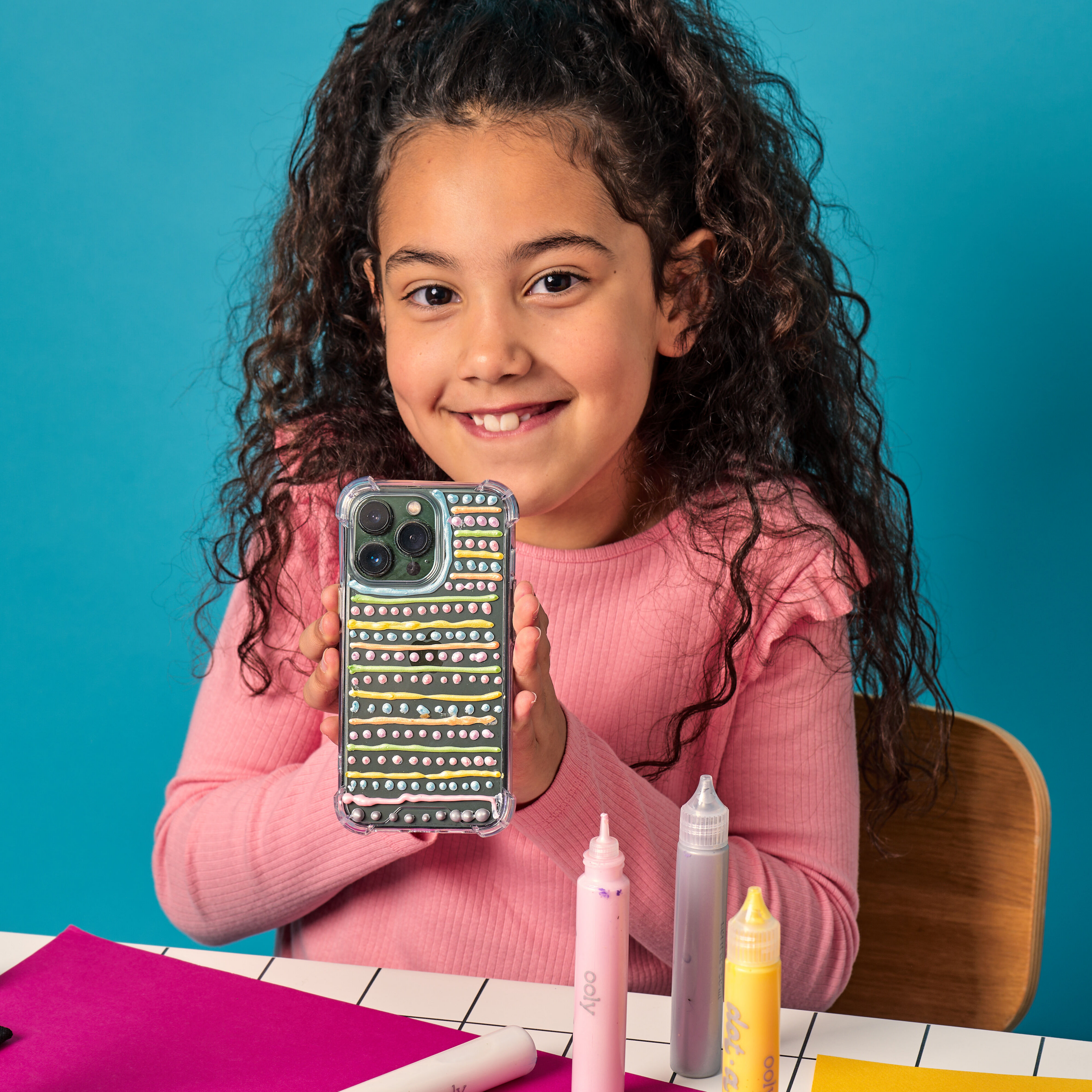 Young girl holding a phone with a colorful case painted with OOLY Dot-a-Lot 3D Dimensional Craft Paint against a blue background