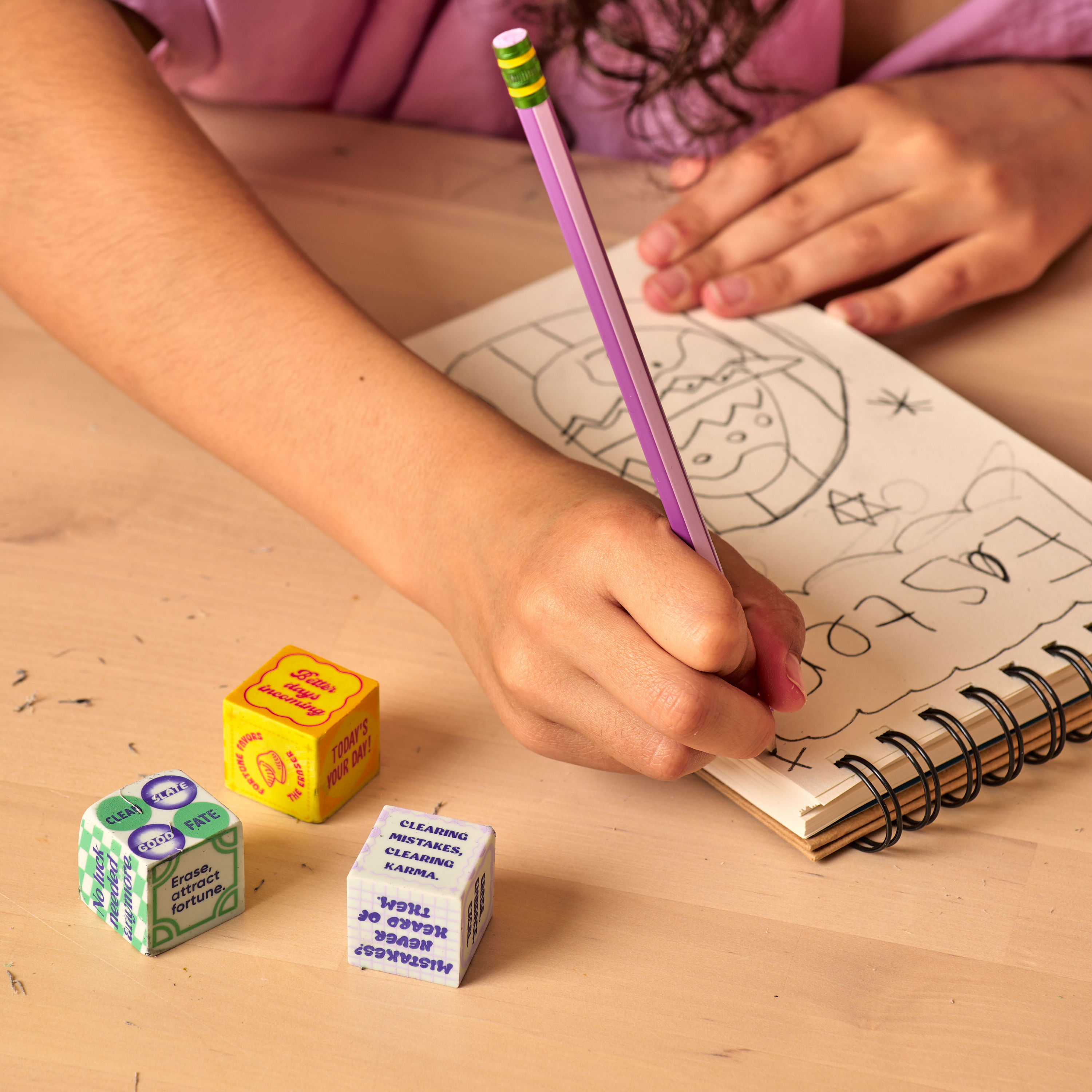 Close up of a girl drawing in a sketchbook with OOLY Feeling Lucky Dice Erasers at her side on a wooden table