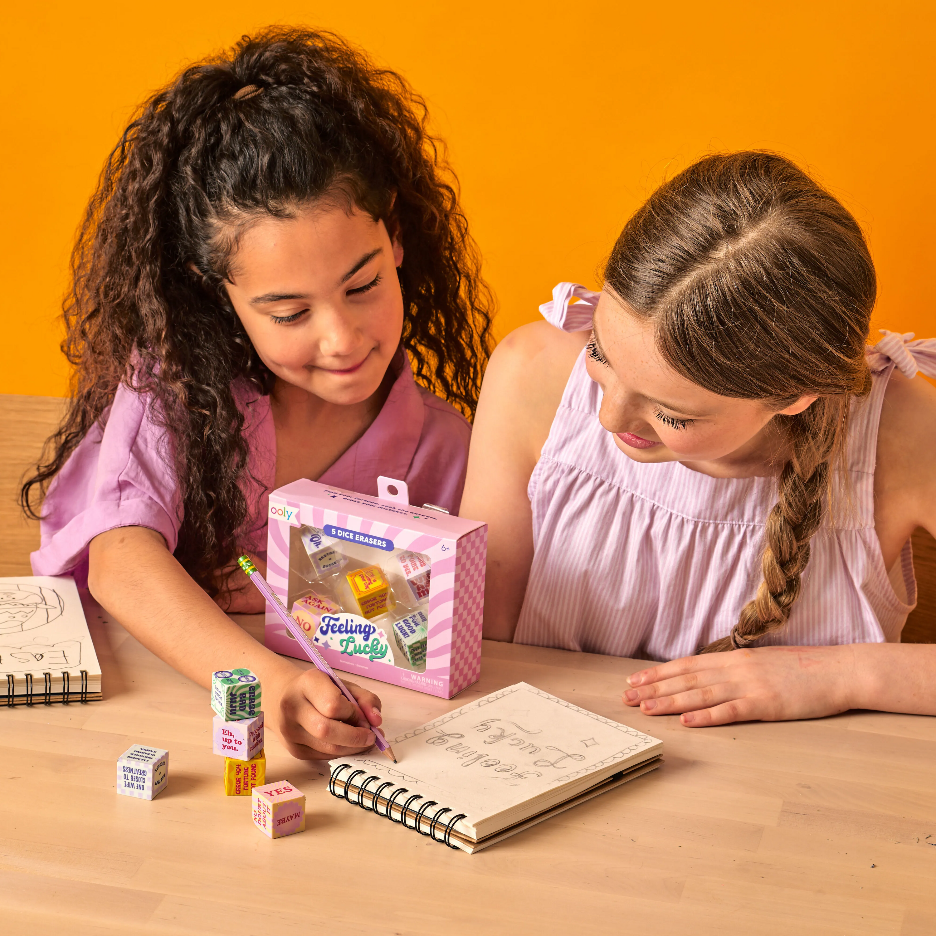 Two girls drawing in a sketchbook with OOLY Feeling Lucky Dice Erasers at their side on a wooden table
