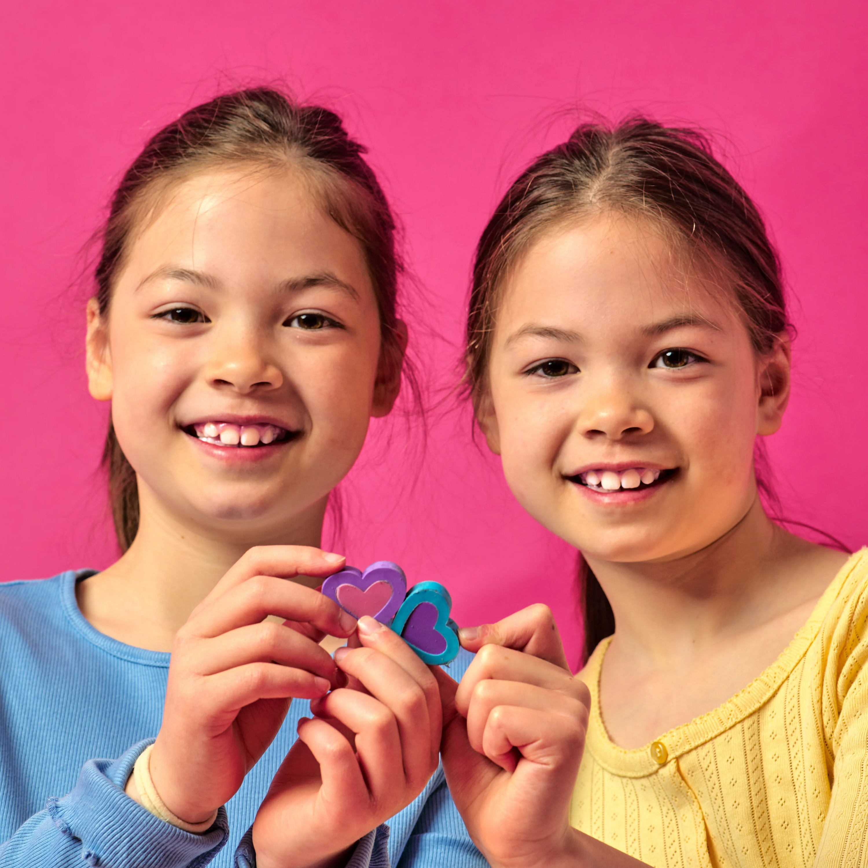 Two girls holding OOLY Hearts in Bloom Puzzle Erasers and smiling against a hot pink background