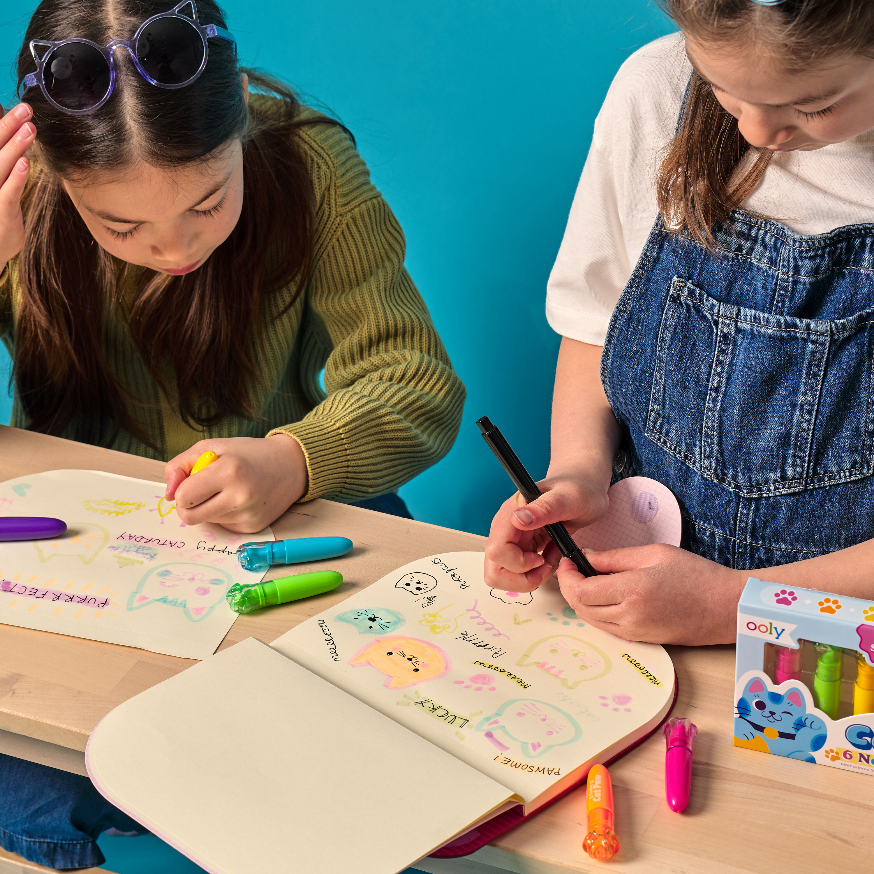 Two girls highlighting cat drawings with OOLY Lucky Cat Paw Scented Neon Highlighters on a wooden table