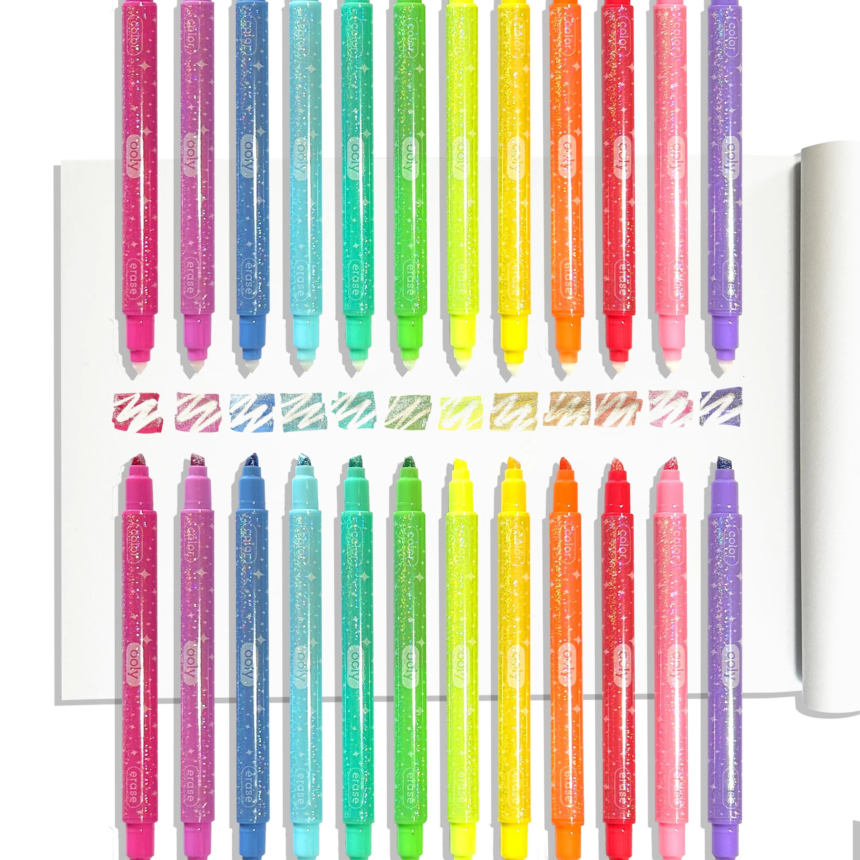 Row of OOLY Make No Mistake! Glittery Erasable Markers uncapped on a white sketchbook showing erasable glitter swatches