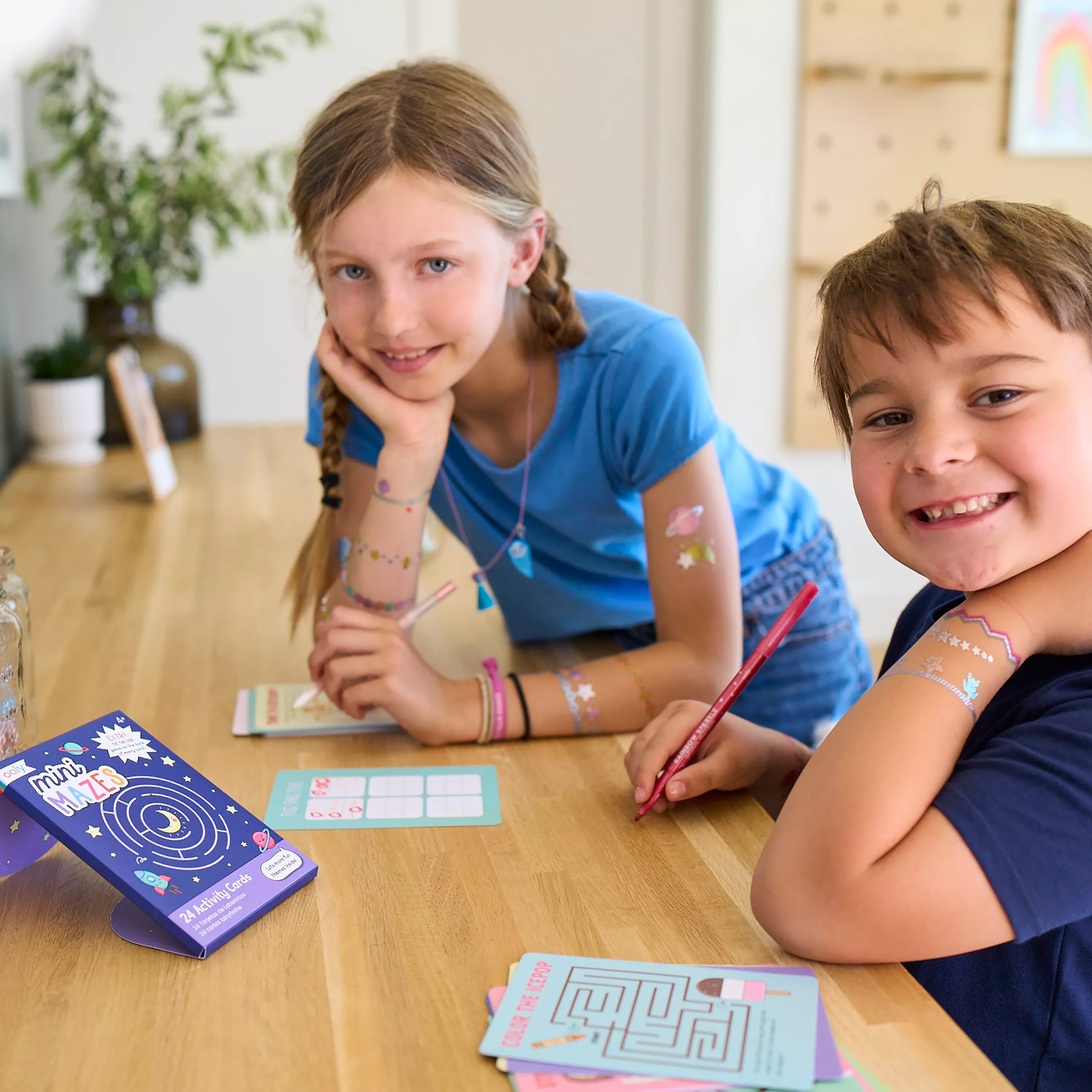 Two kids playing games from a set of OOLY Mini Mazes Activity Cards and smiling at camera