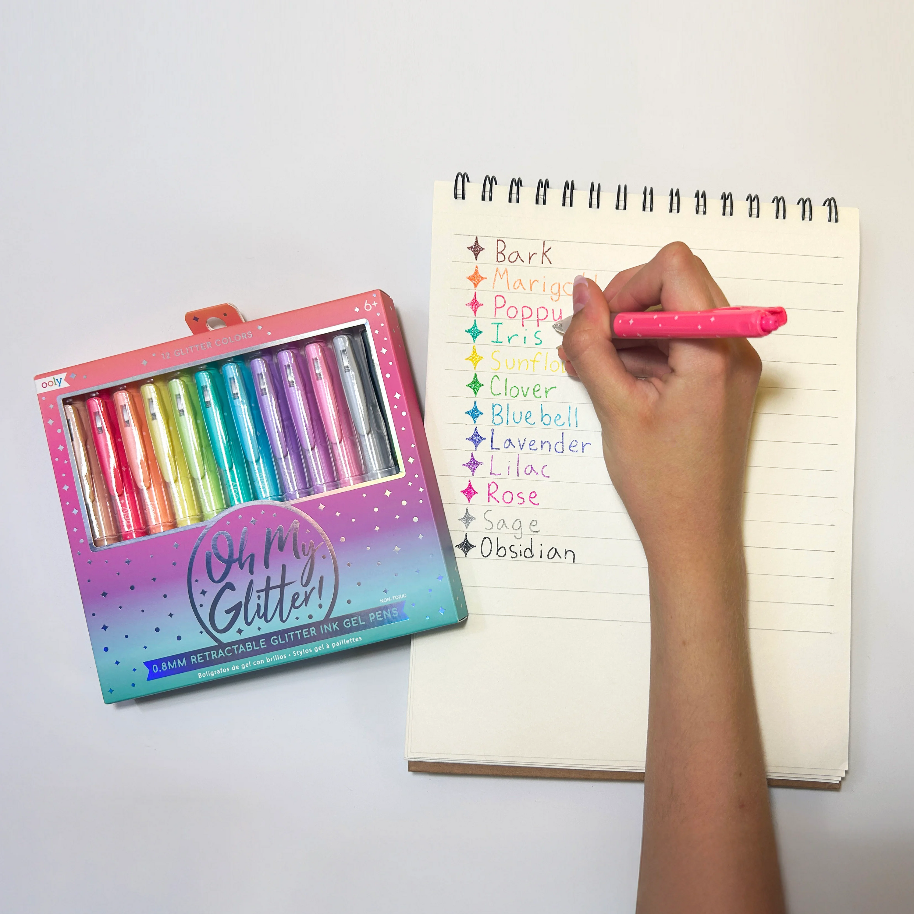 Hand writing on notepad different colored swatches with set of OOLY Oh My Glitter! Retractable Gel Pens nearby
