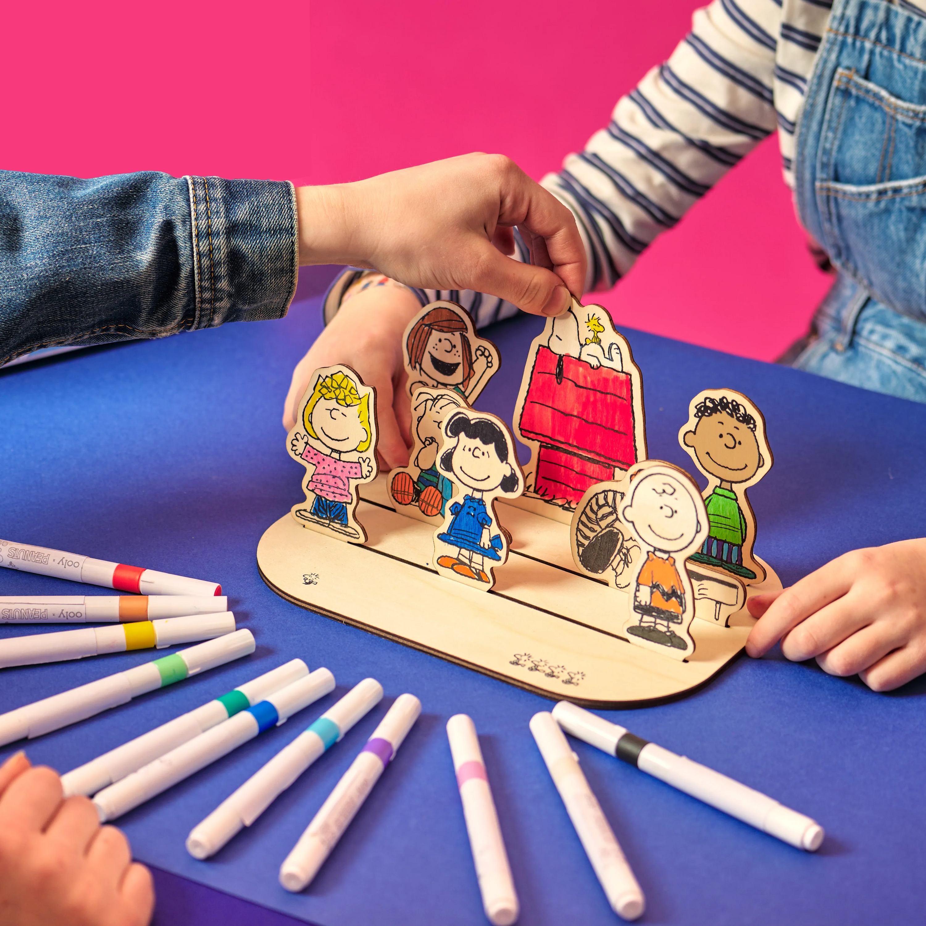 Two people setting up the OOLY x Peanuts Color-In Wooden Scene with paint markers