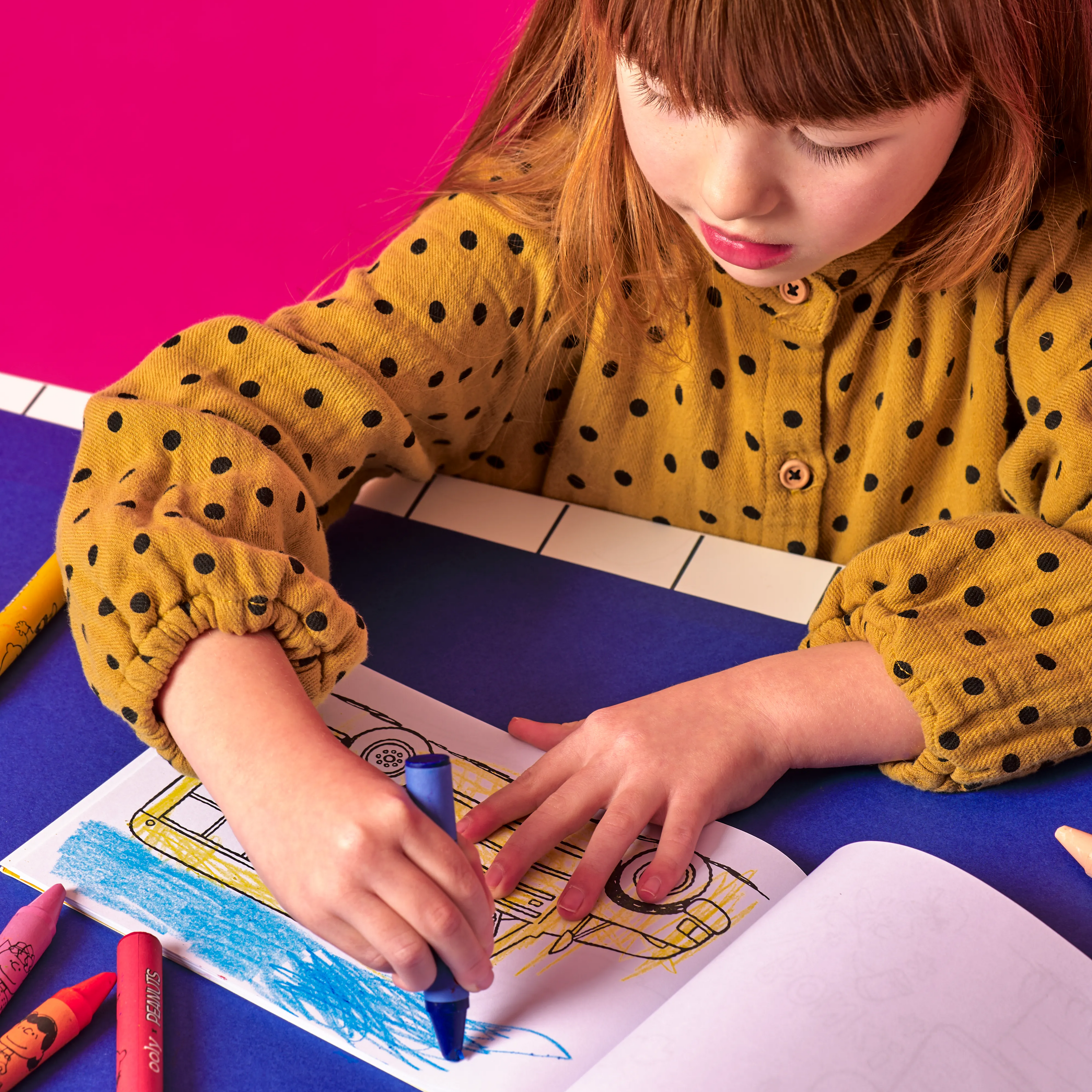 Girl coloring a school bus coloring page with a blue crayon from the OOLY x Peanuts Snoopy School Coloring Travel Kit