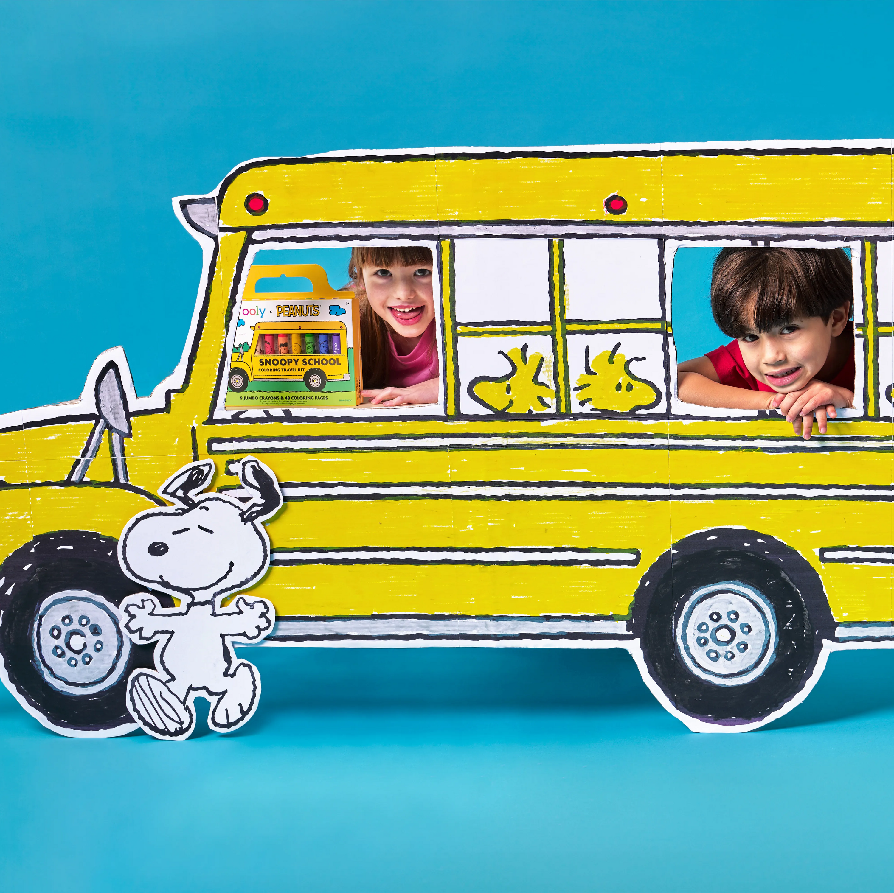 Two kids sitting in a colored prop school bus with a Snoopy cutout holding up an OOLY x Peanuts Snoopy School Coloring Travel Kit