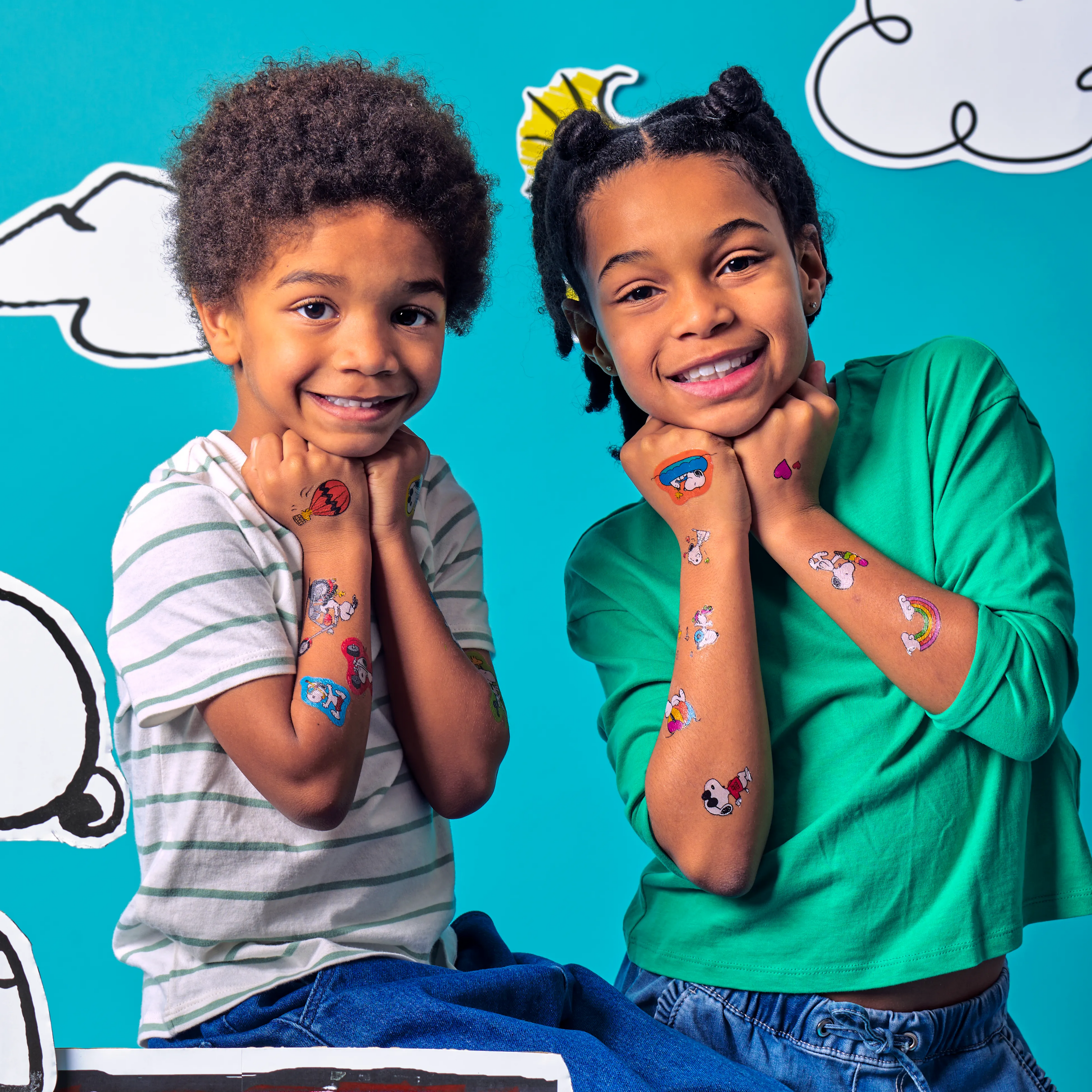 Two kids against a cloudy blue background showing off temporary tattoos on their arms from an OOLY x Peanuts Temporary Tattoos set