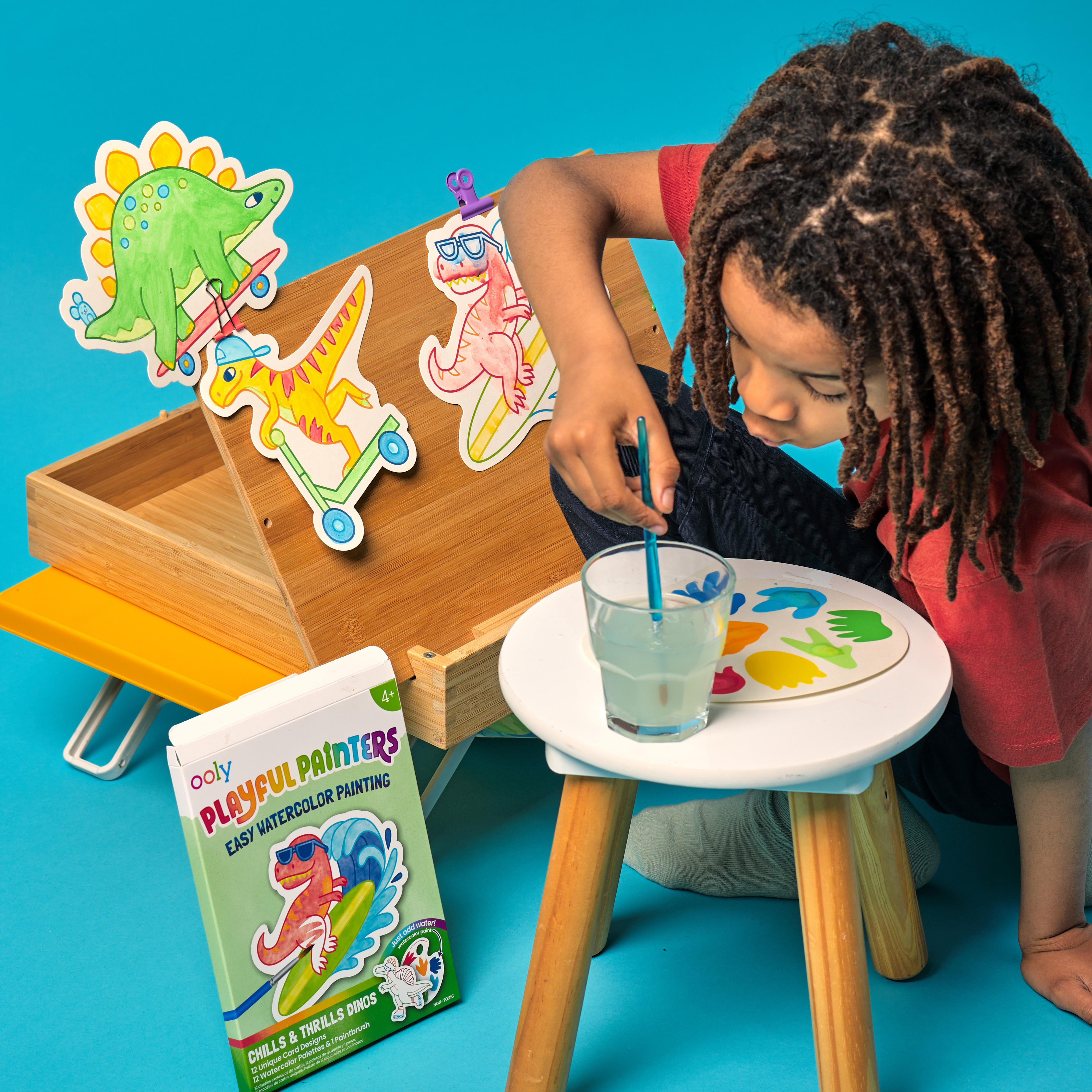 Kid painting with an OOLY Chills and Thrills Dinos Playful Painters Easy Watercolor Painting Kit on a blue background