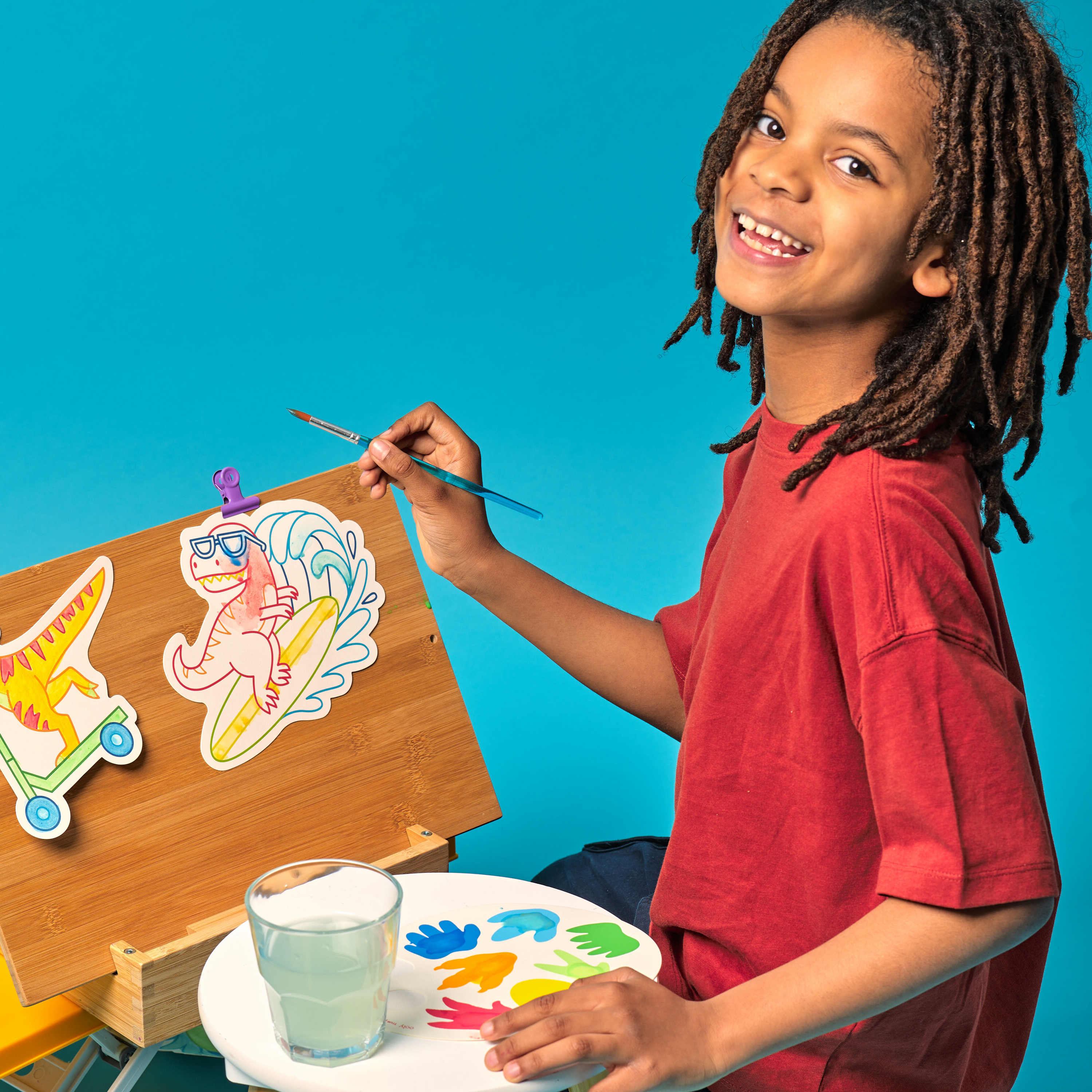Kid smiling while painting dinosaur paint cards from the OOLY Chills and Thrills Dinos Playful Painters Easy Watercolor Painting Kit on a blue background