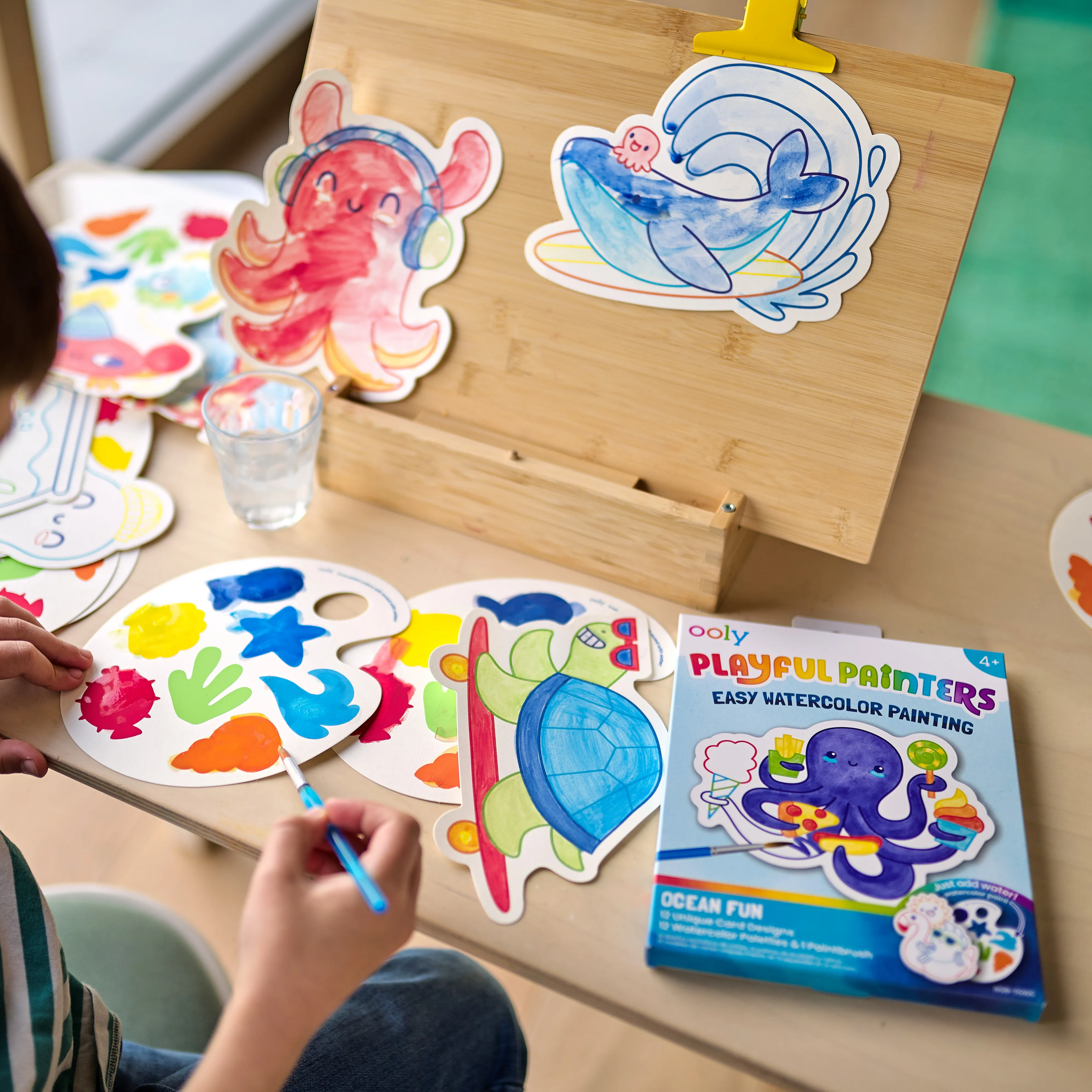 Kid with paint cards and palettes from the OOLY Ocean Fun Playful Painters Easy Watercolor Painting Kit on a wooden easel