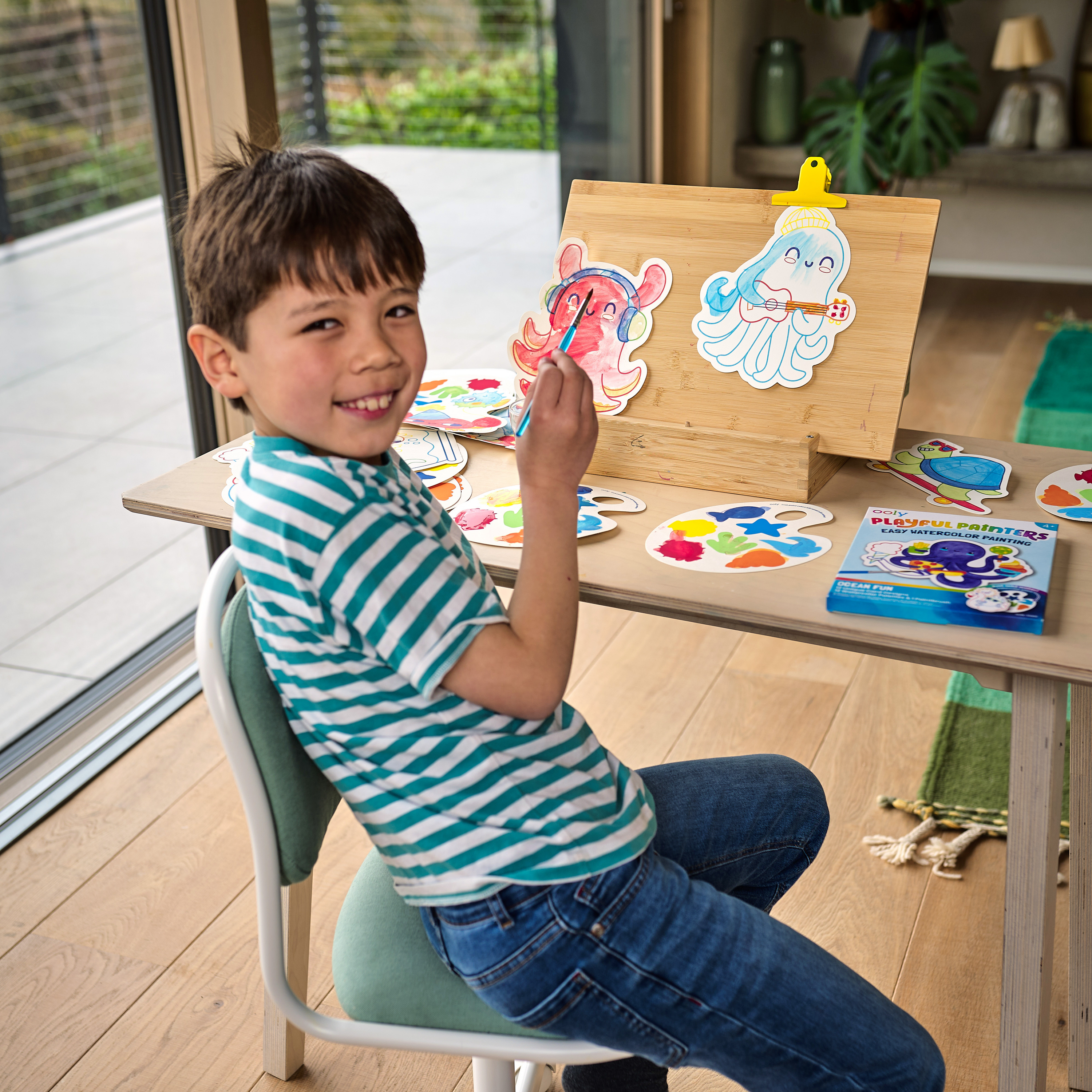 Kid smiling while painting with OOLY Ocean Fun Playful Painters Easy Watercolor Painting Kit on a wooden easel.