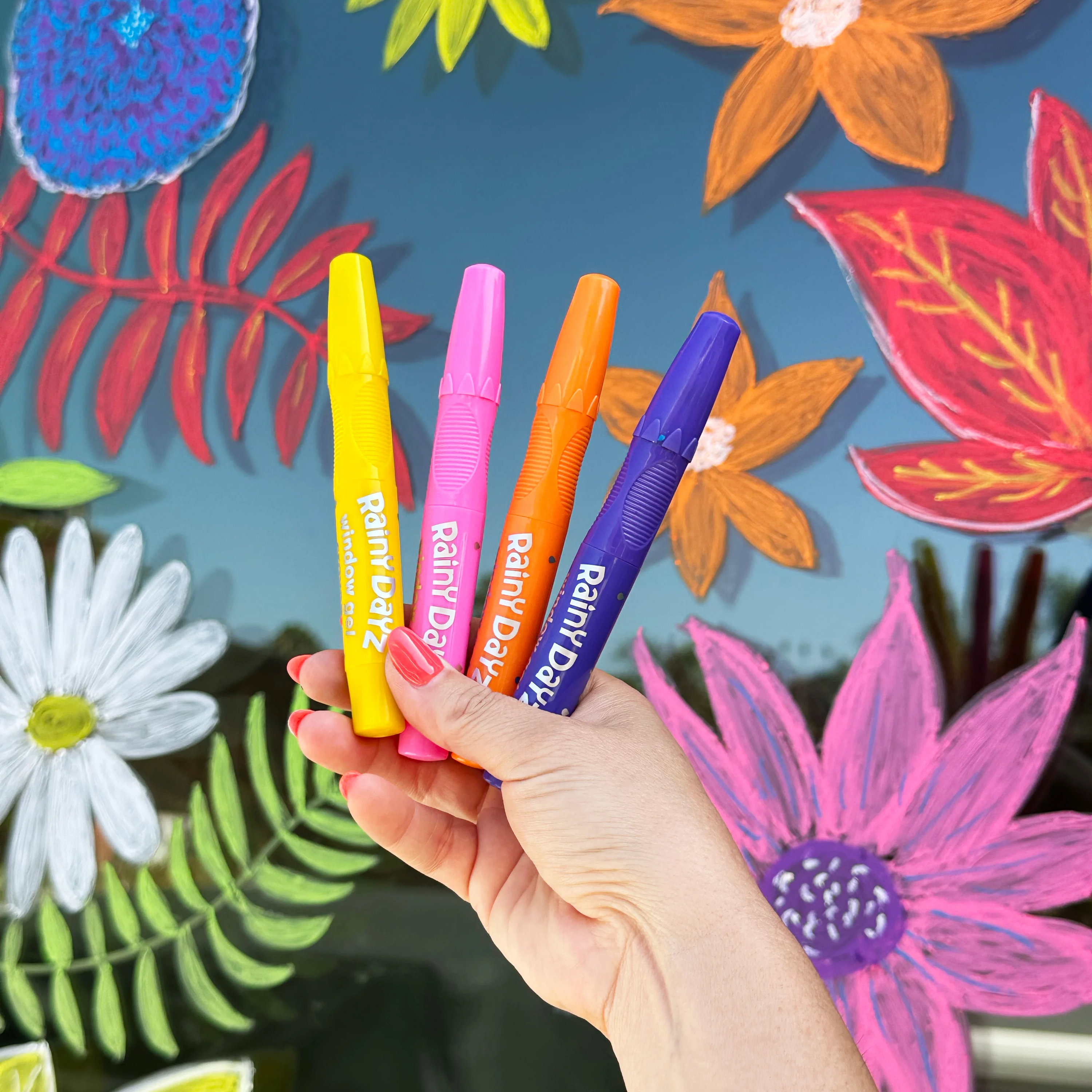 Woman's hand holding four OOLY Rainy Dayz Window Gel Crayons in front of a window with colorful flower drawings