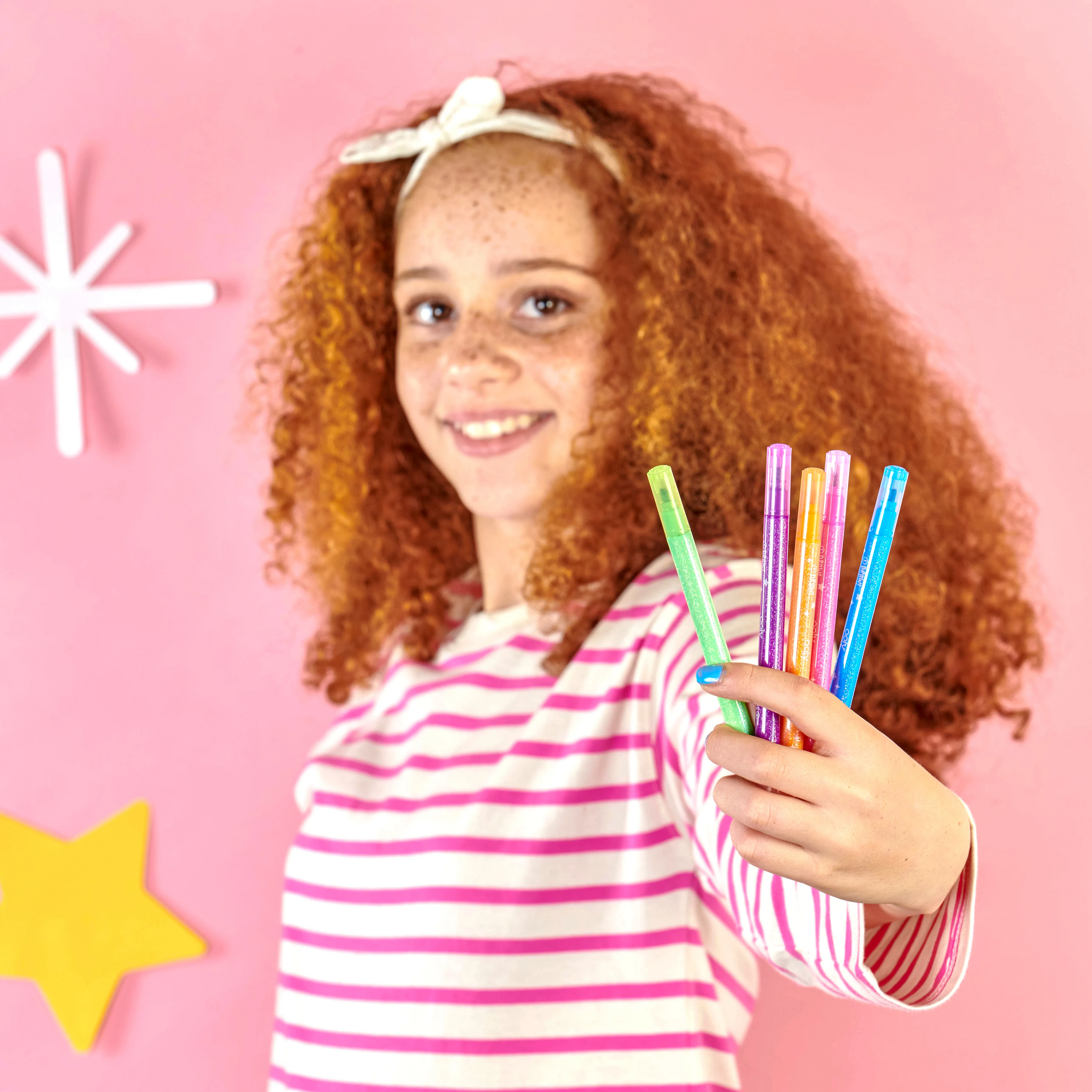 Girl holding up five OOLY Switch-Eroo Sparkle Glittery Color Changing Markers with stars and pink background