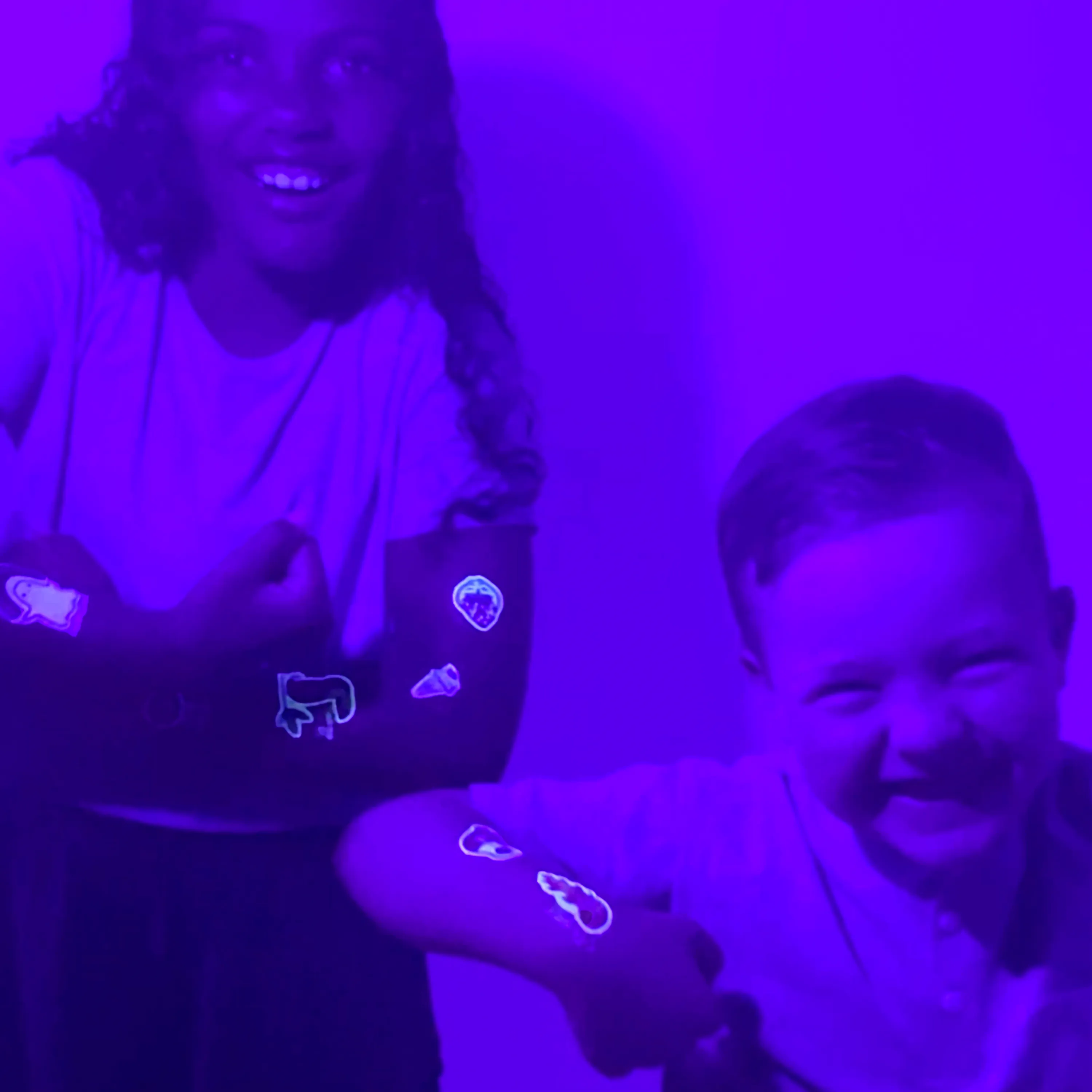 Two kids in black light room with OOLY Tattoo-Palooza Glow in the Dark Temporary Tattoos on their arms
