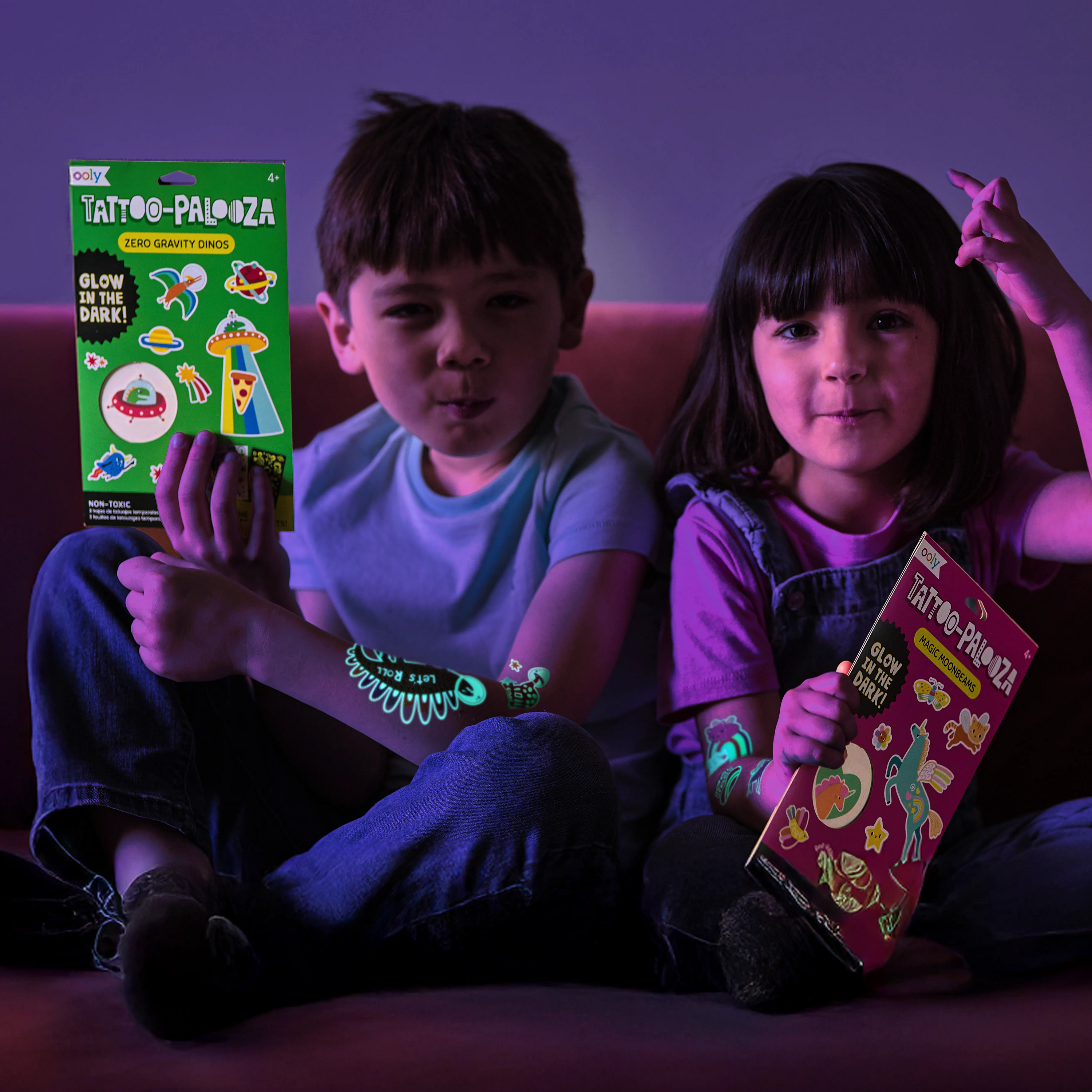 Two kids in darkly lit room holding sets of OOLY Tattoo-Palooza Glow in the Dark Temporary Tattoos