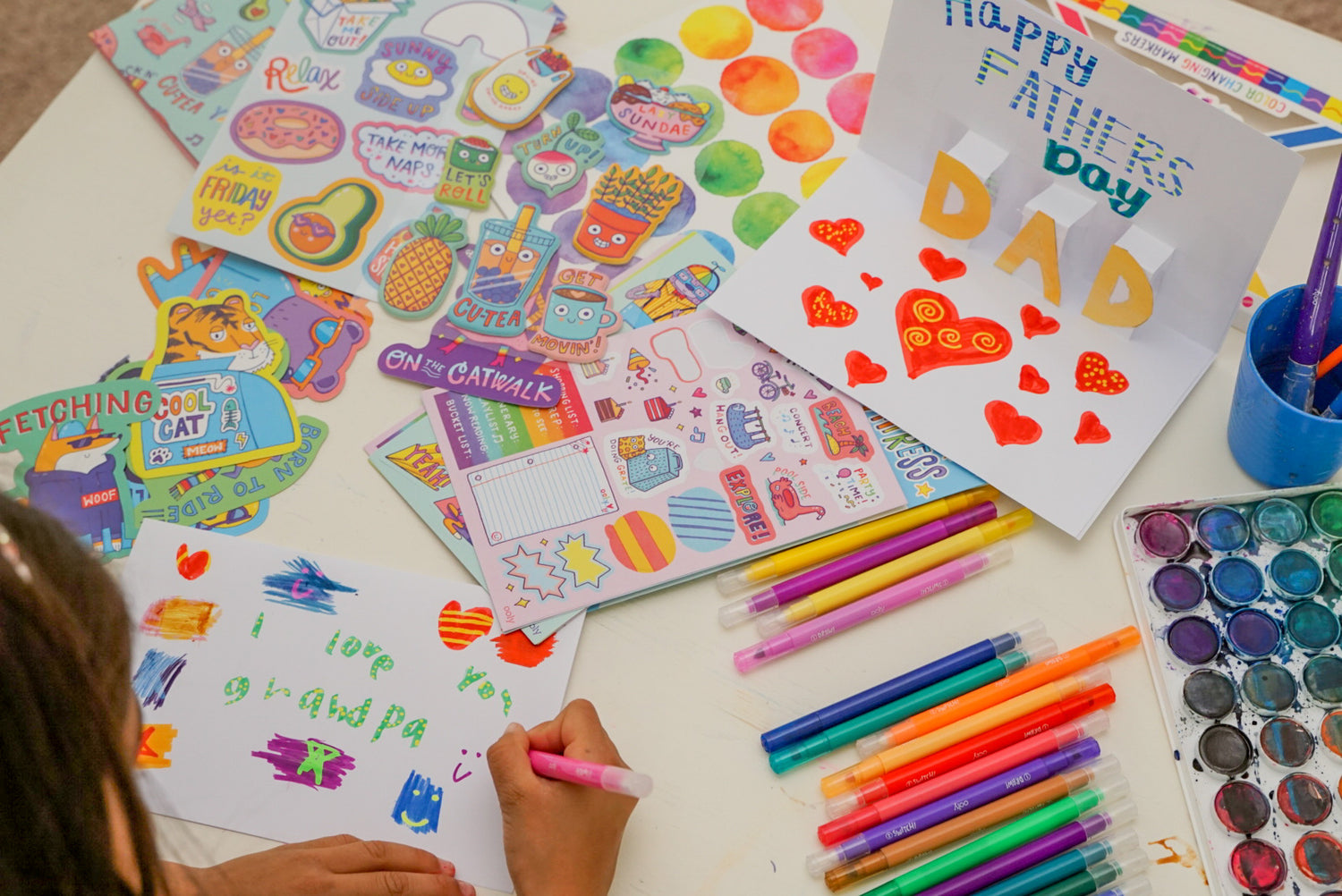 Kid using markers to write i love you grandpa on handmade card with arts and crafts supplies on table