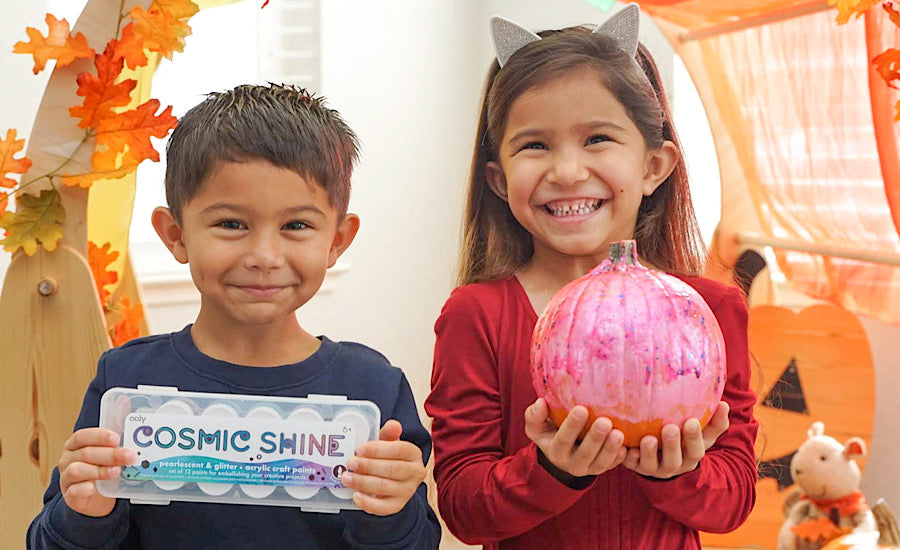 two smiling kids holding paint and a pumpkin surrounded by fall decor