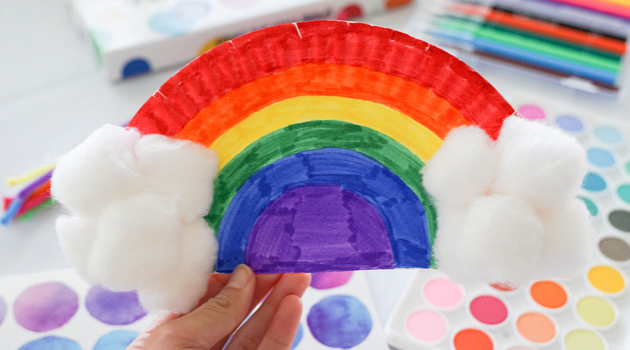 hand holding rainbow made out of paper plate with art supplies in background