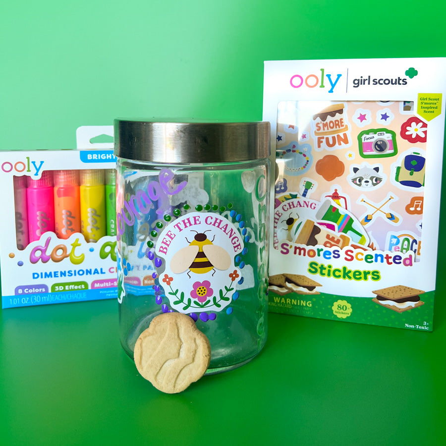Glass friendship jar decorated with OOLY x Girl Scouts S’mores scented stickers and colorful paint details, filled with paper affirmations and Girl Scout cookies on a green background. 