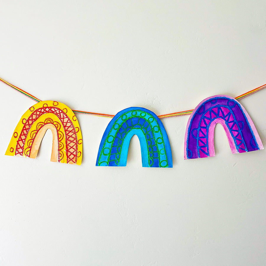 Three colorful rainbow shapes with intricate wax crayon designs in red, blue, and green, painted with vibrant watercolor hanging on a white wall with string. OOLY Craft blog.