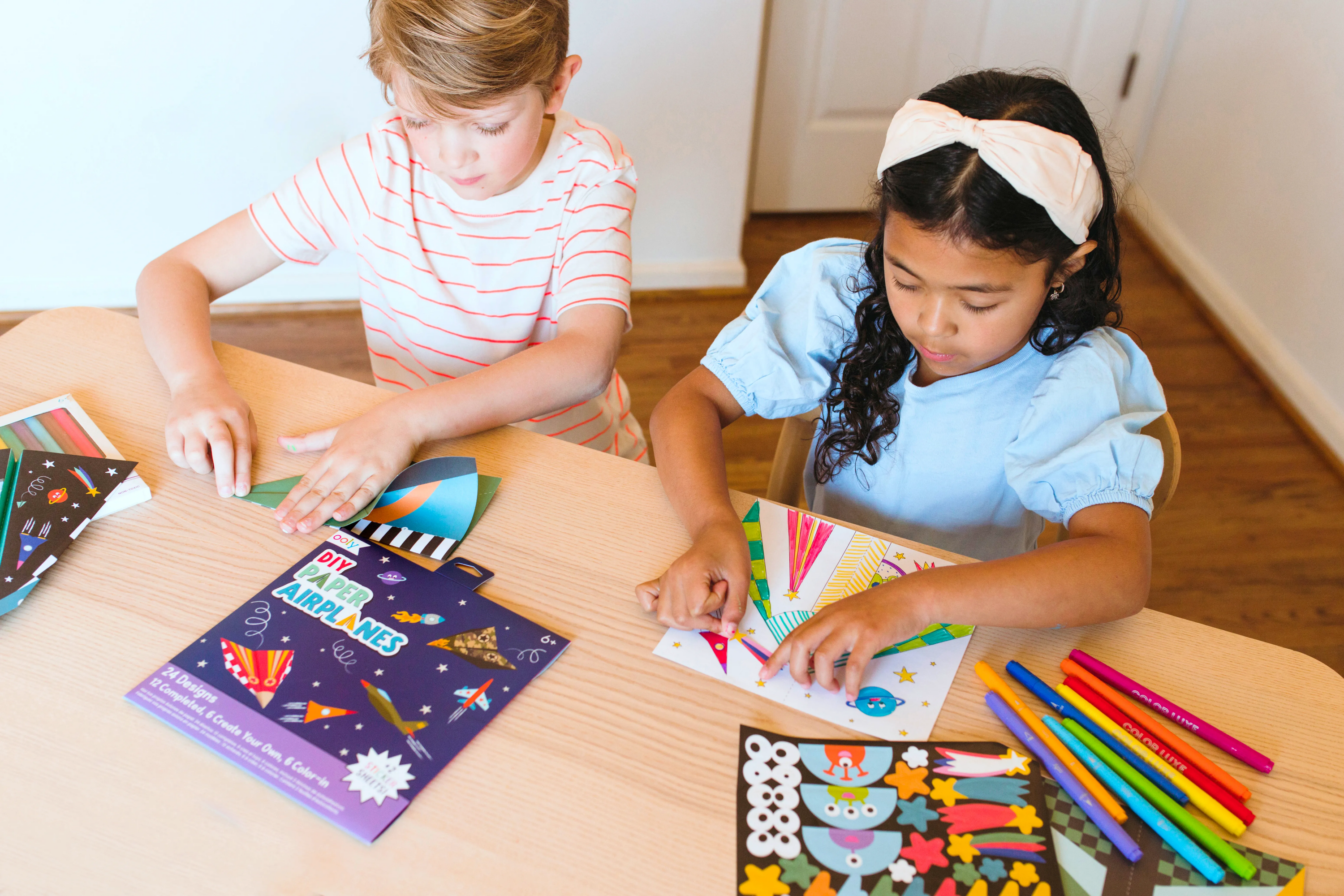 Two kids folding and applying stickers to OOLY DIY Paper Airplanes Activity Kit