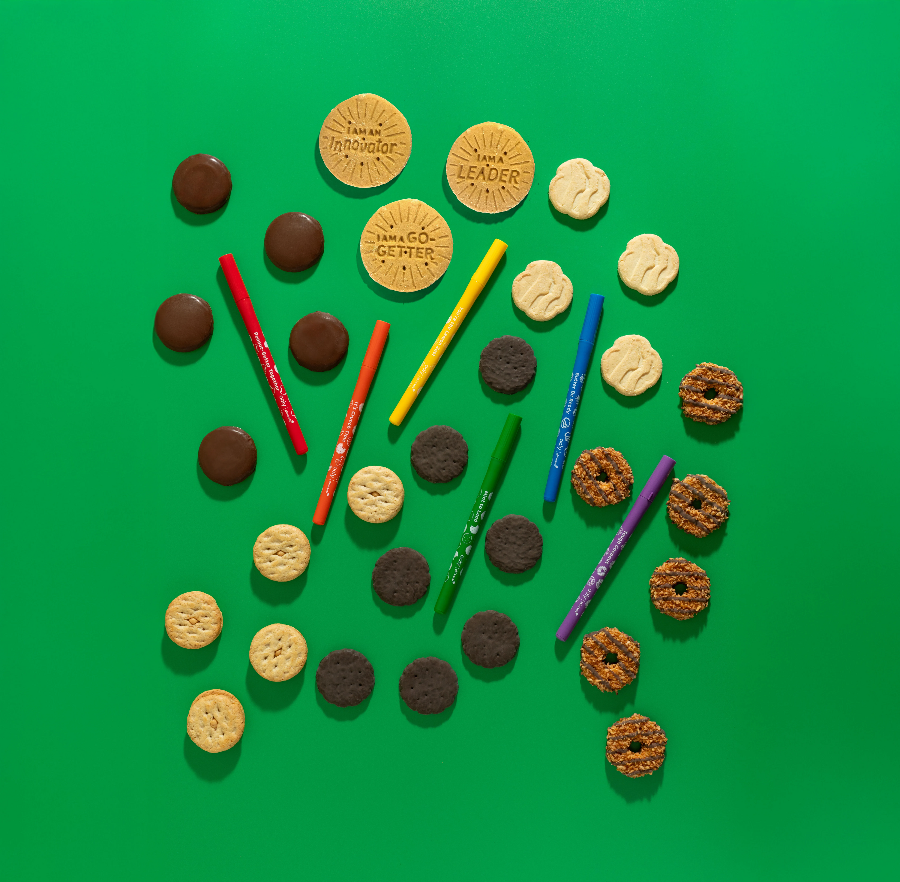 OOLY x Girl Scouts Cookie Scented Gel Pens and Girl Scout cookies spread out in a pattern on a green background