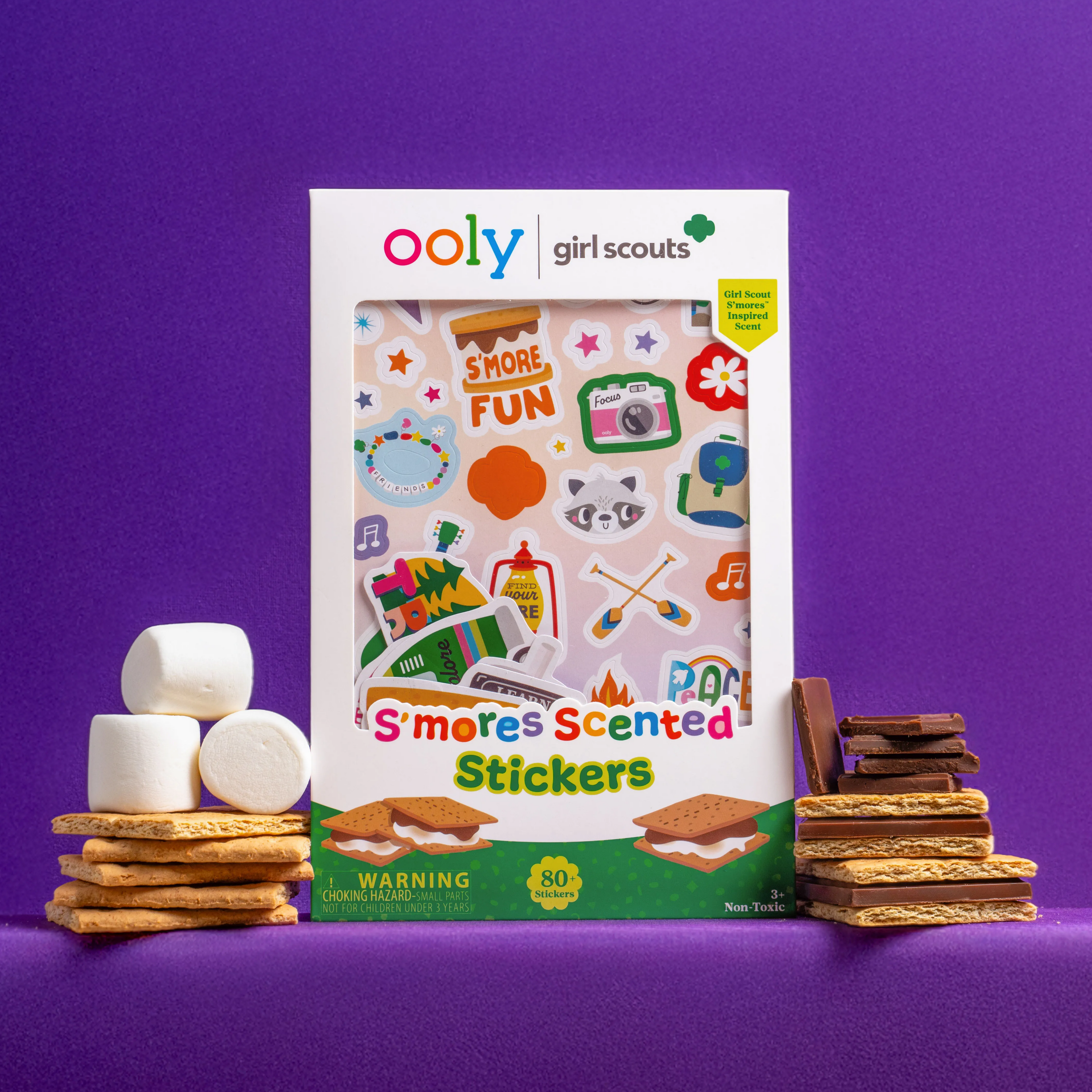 A set of OOLY x Girl Scouts S’more Scented Stickers surrounded by s'mores ingredients on a purple background
