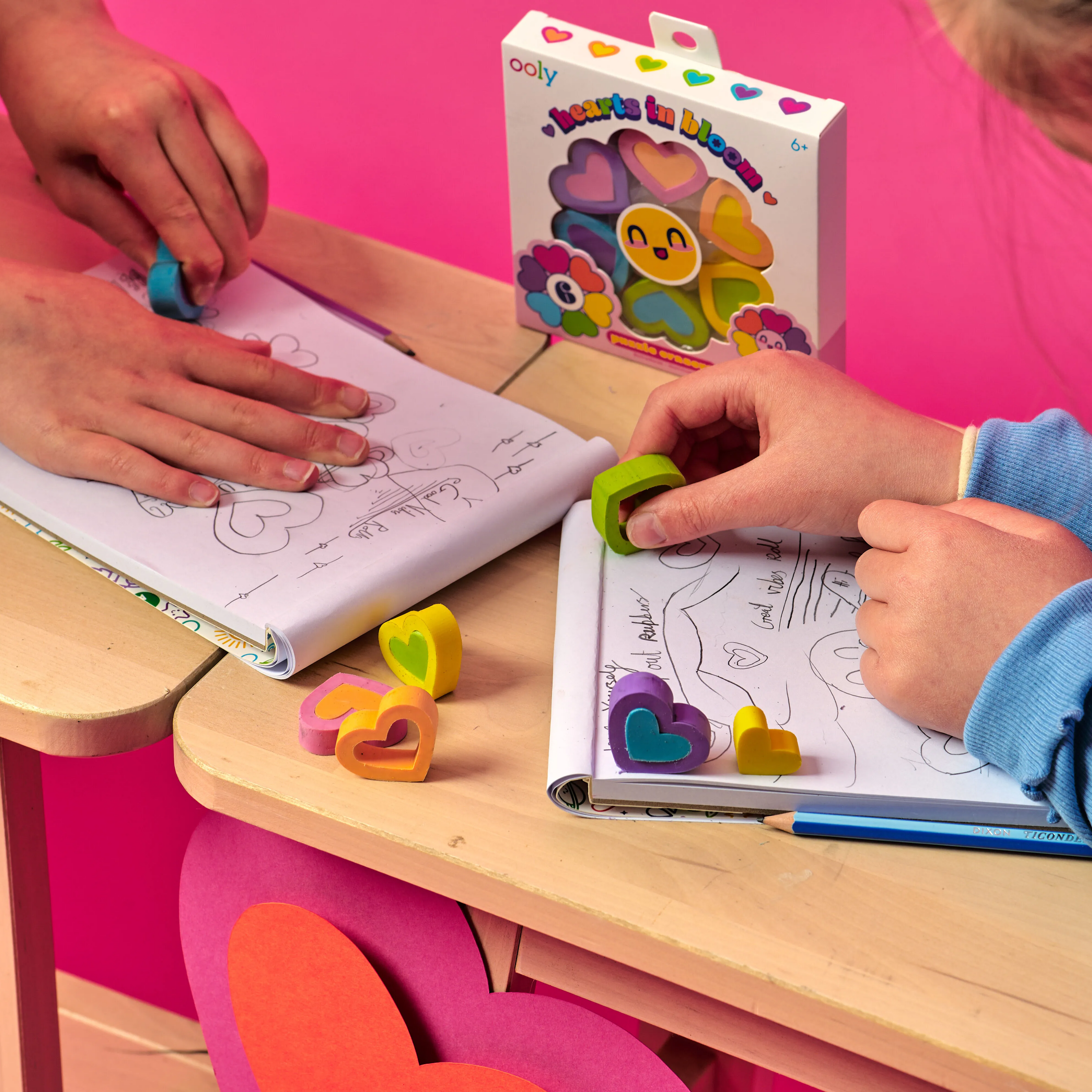 Two people erasing drawings in white sketchbooks with OOLY Hearts in Bloom Puzzle Erasers on wooden tables