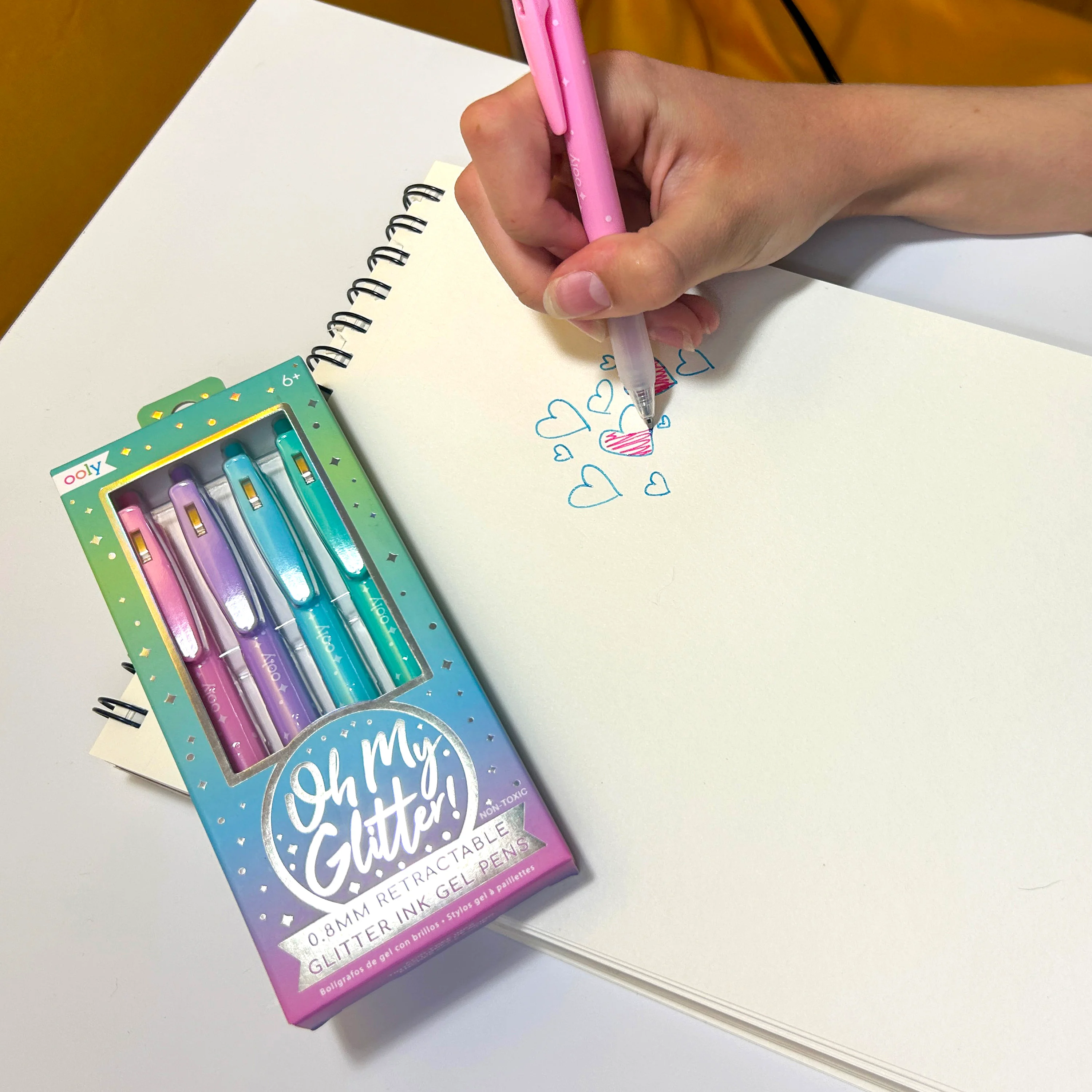 Hand coloring in blue hearts with a pink OOLY Oh My Glitter! Retractable Gel Pen on a sketchpad