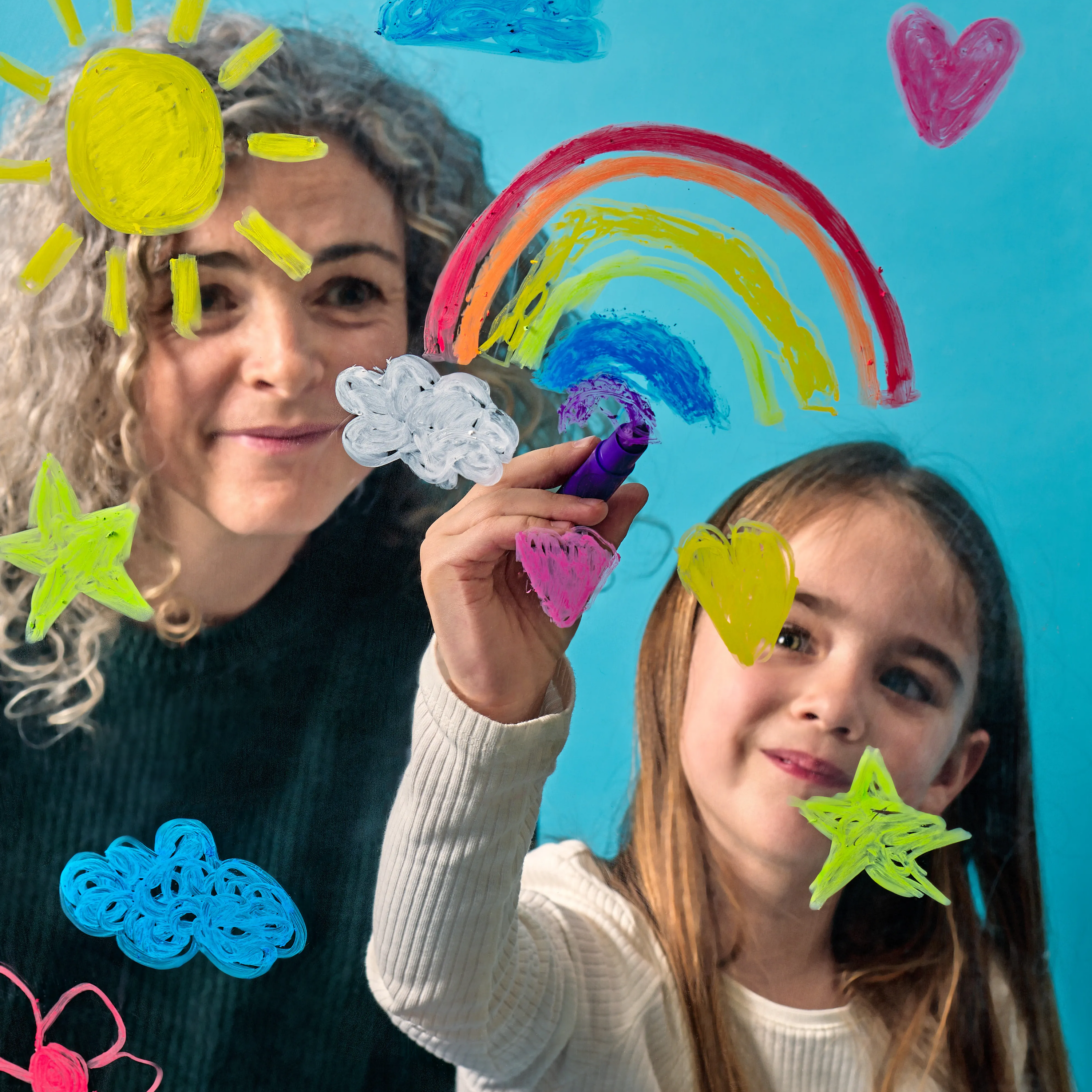 Girl coloring rainbows, hearts and clouds on a window with OOLY Rainy Dayz Window Gel Crayons while a woman watches