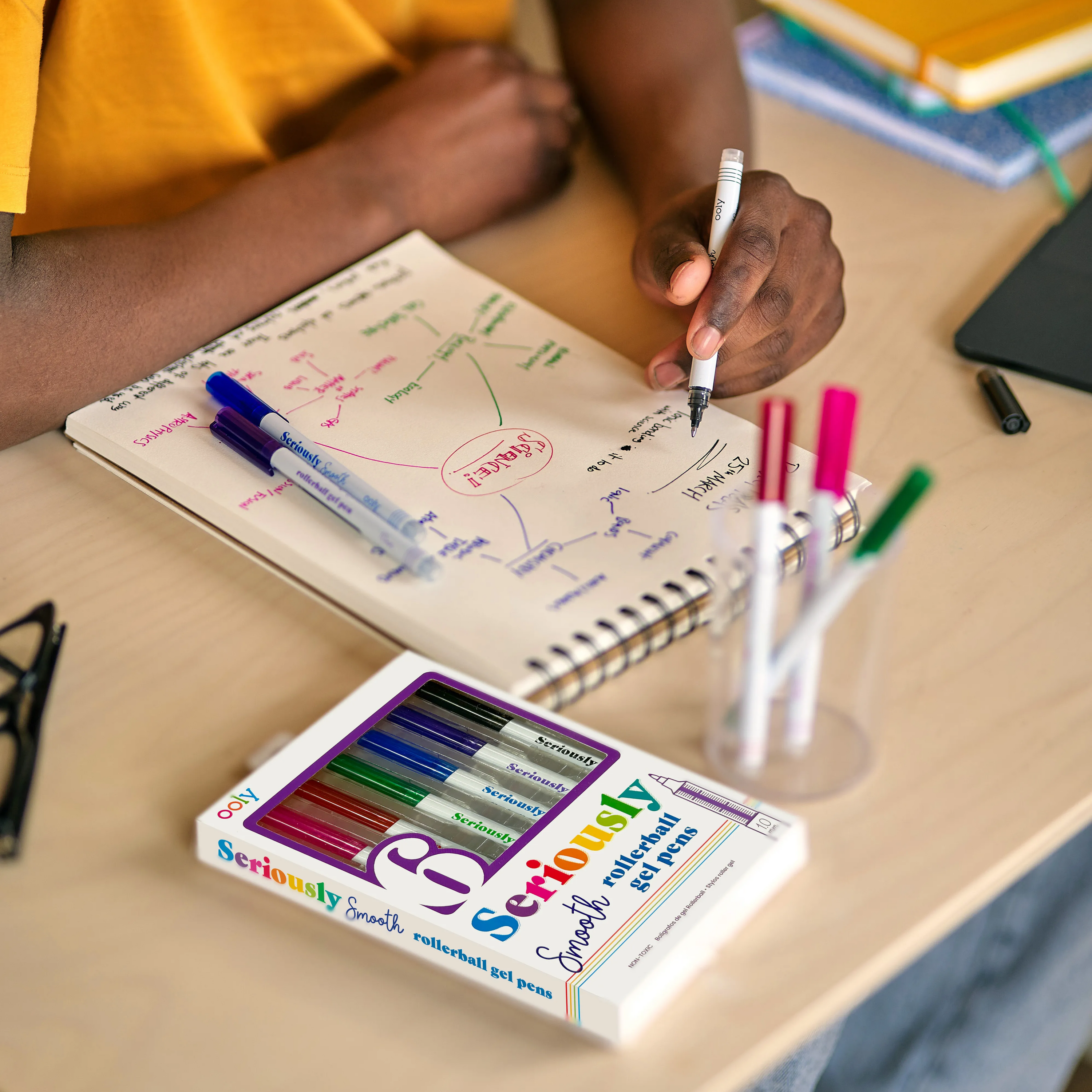 Kid writing a brainstorming guide in a white skethbook with OOLY Seriously Smooth Rollerball Gel Pens on a wooden desk