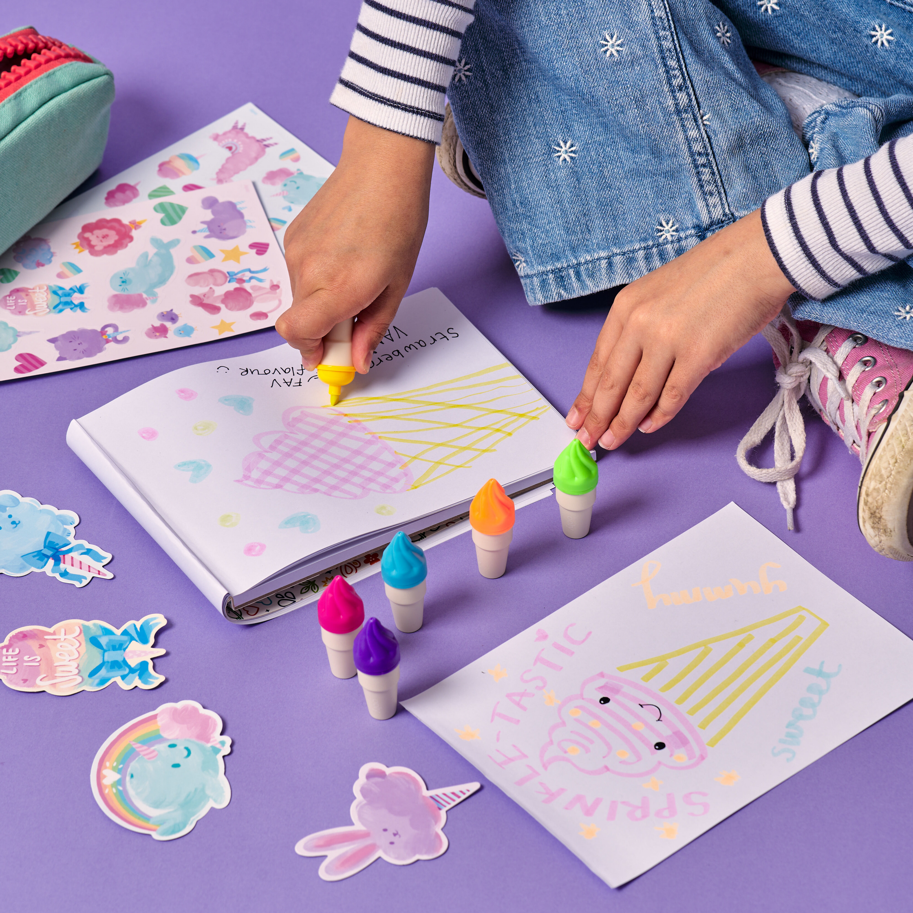 Close up of a girl drawing with OOLY Sweet Scoops Scented Neon Highlighters in a white sketchbook on a purple background
