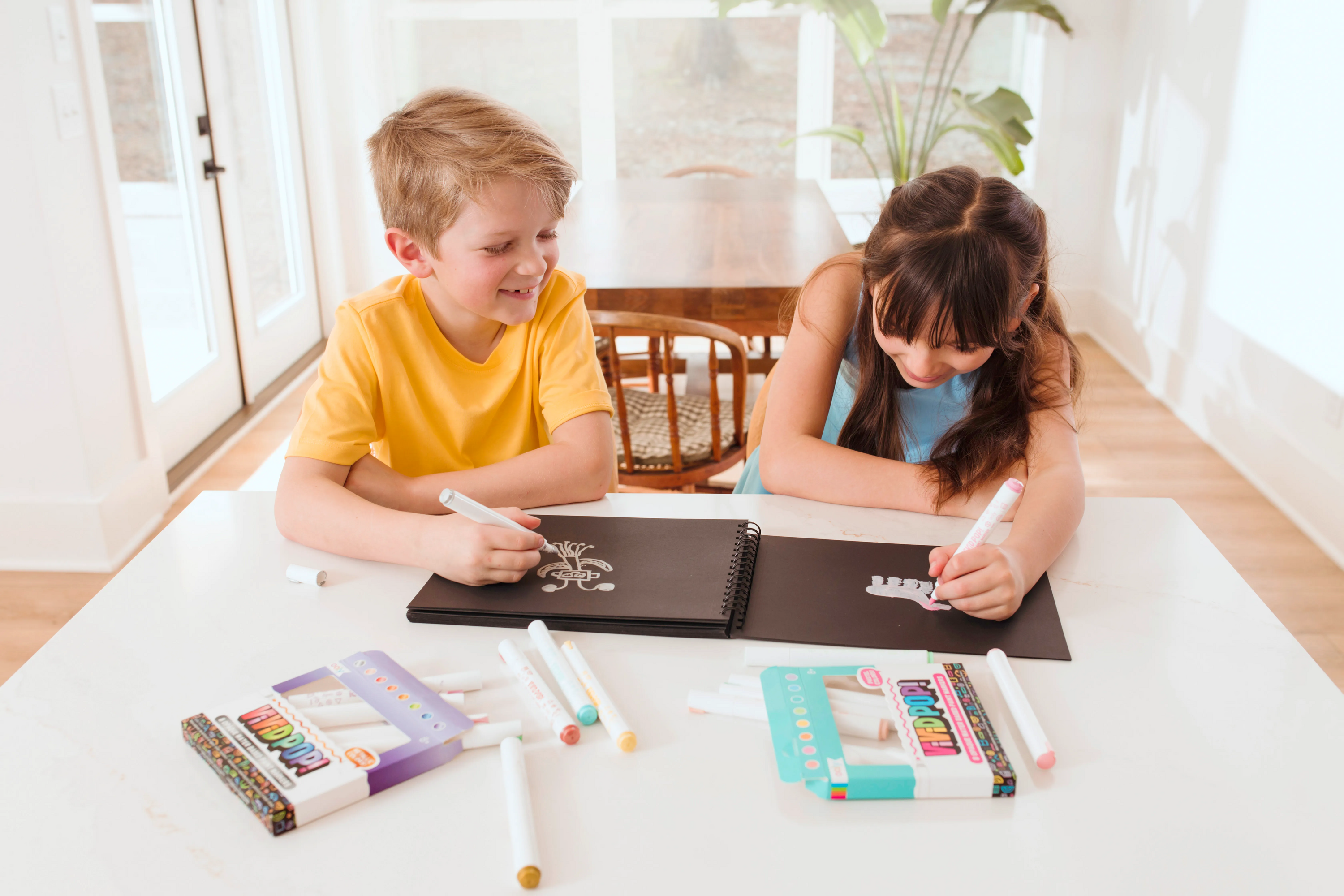 Two kids coloring on black paper pad with OOLY Vivid Pop! Paint Markers wide shot
